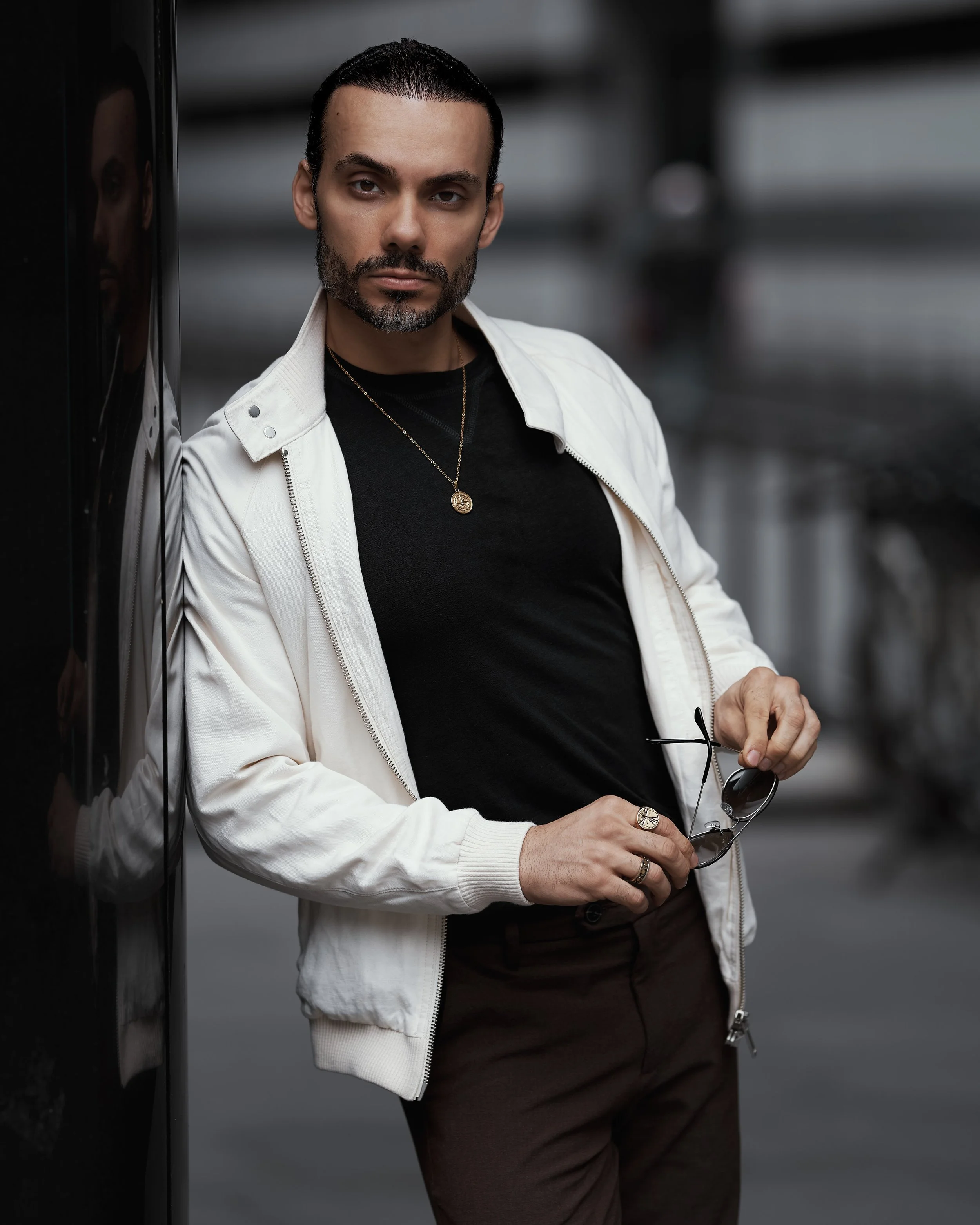 Junge Mann mit dunklem Haar, Bart, schwarze T-Shirt, weisjem Mantel, trägt eine Halskette und eine Uhr, hält eine Sonnenbrille, steht vor einem schwarzen Hintergrund.