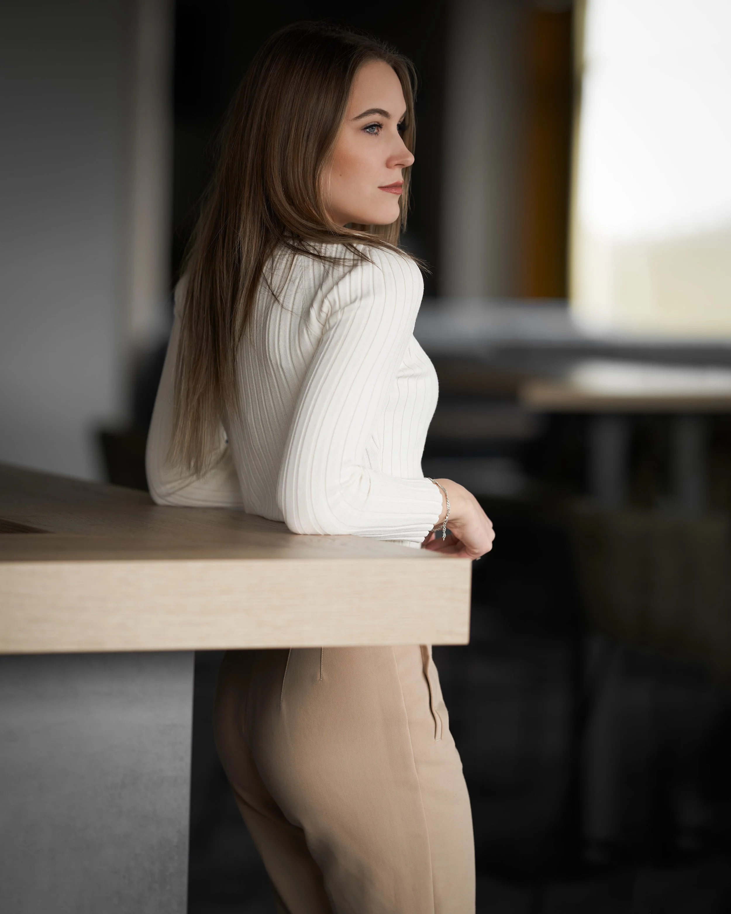 Nahaufnahme einer jungen Frau mit braunen Haaren, die nach rechts schaut, in einem hellen Oberteil, an einem Tisch sitzend, in einer modernen, dunklen Raumumgebung.