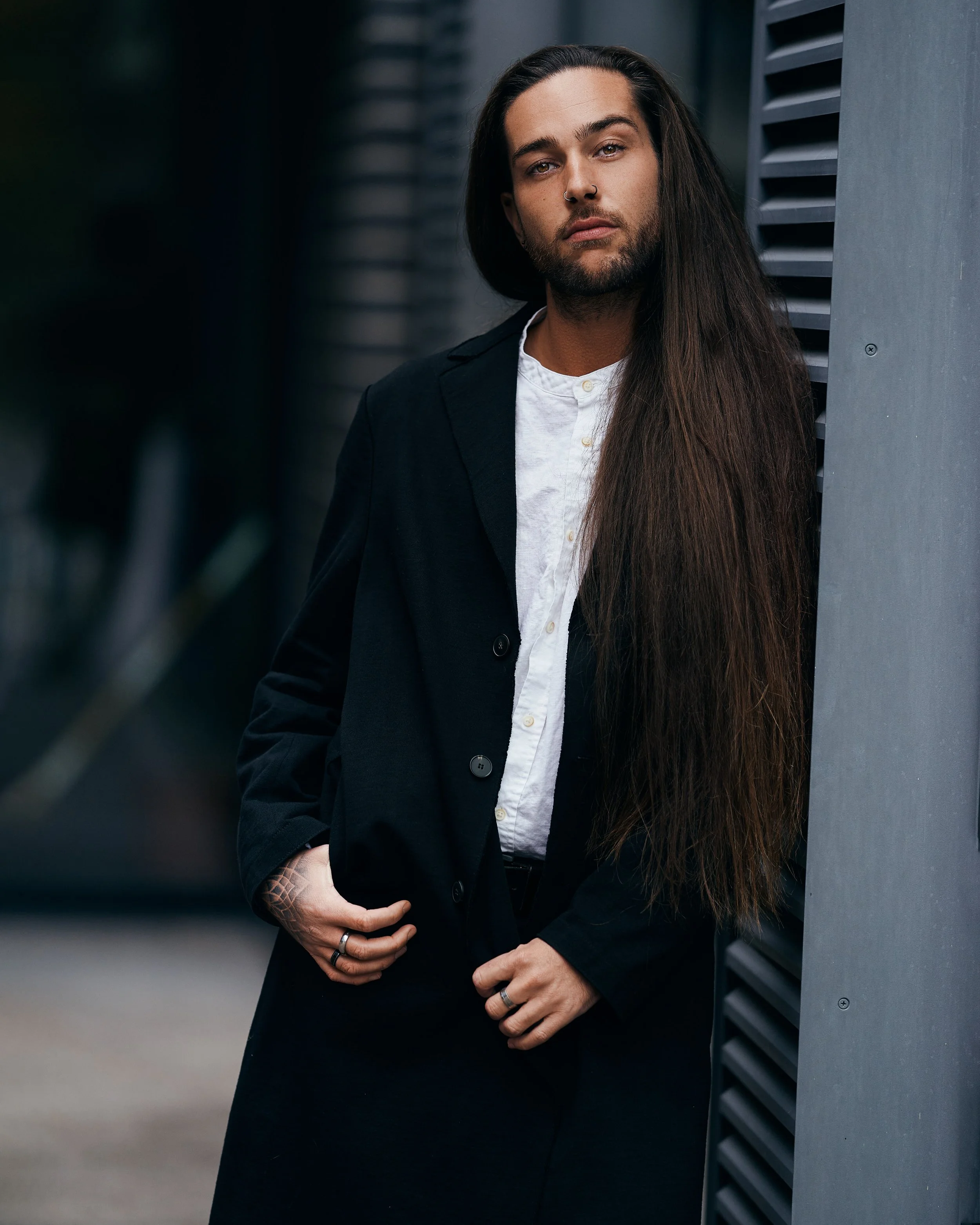 Ein junger Mann mit dunklen, langen Haaren, tätowierten Händen, Nase und Ohrpiercing, trägt einen schwarzen Mantel über weißem Hemd und posiert vor einer grauen Gebäudewand.