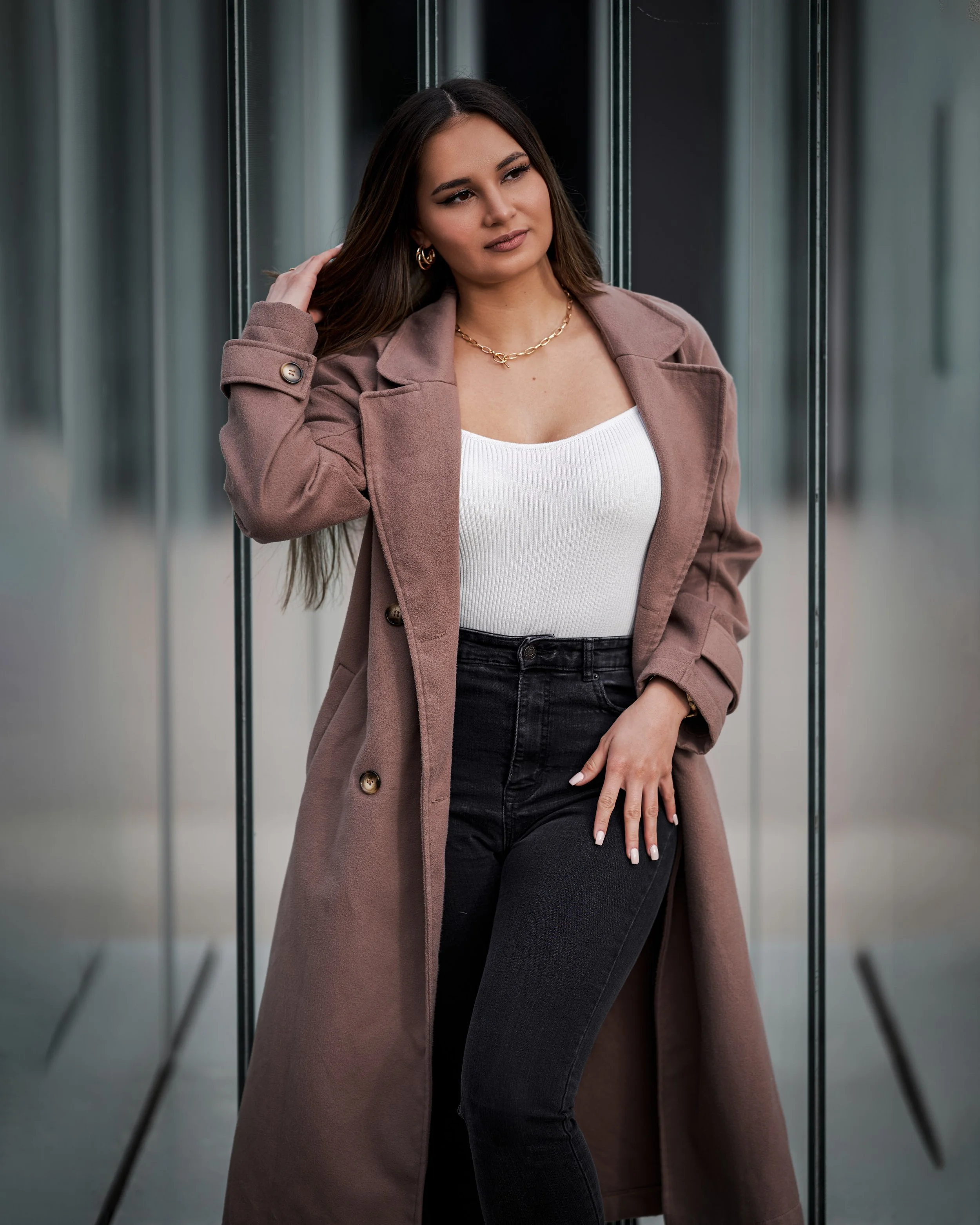 Eine junge Frau mit langen braunen Haaren trägt eine rosa oder braune Jacke, eine weiße Oberteil, schwarze Jeans, Goldschmuck und posiert vor einem spiegelnden Hintergrund.