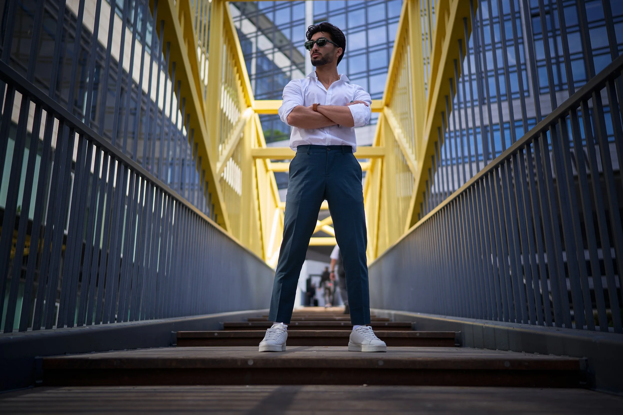 Junger Mann mit dunklem Haar, Bart, Sonnenbrille, weißem Hemd, blauer Jeans, weißen Turnschuhen steht auf einer Brücke zwischen Metallgeländern in einem modernen Gebäudekomplex.