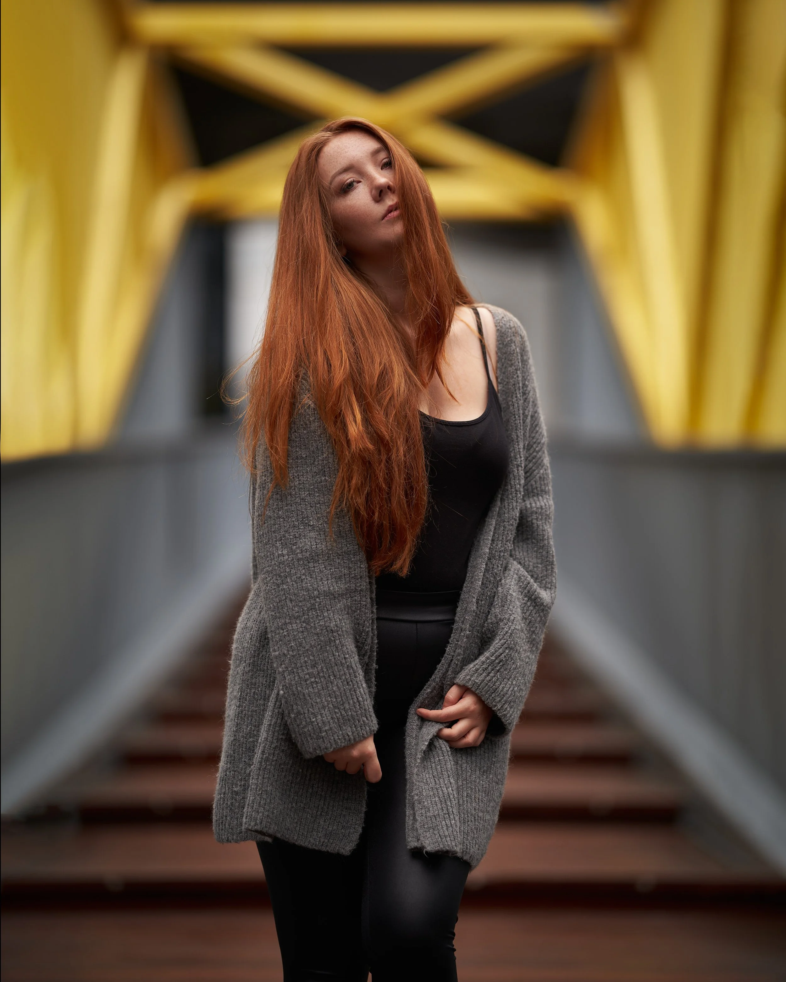 Junge Frau mit roten Haaren, dunklem Oberteil und grauem Mantel, steht vor einer Geländerbrücke mit gelb-schwarzem Geländer, Blick leicht seitlich, nahe Kamera, verschwommener Hintergrund.