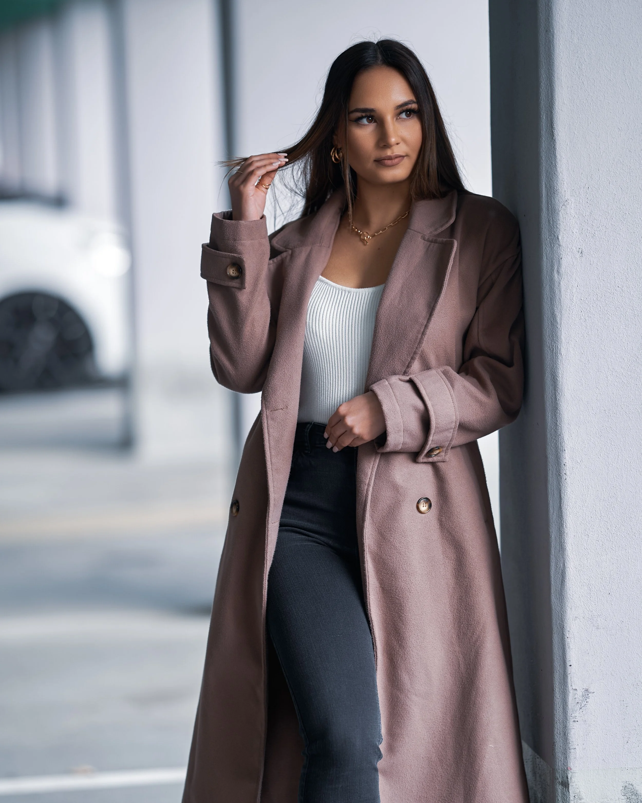 Junge Frau steht an einer weißen Wand, trägt einen beigen Mantel und schwarze Jeans, hat dunkles, schulterlanges Haar, trägt Goldschmuck und schaut nach rechts.