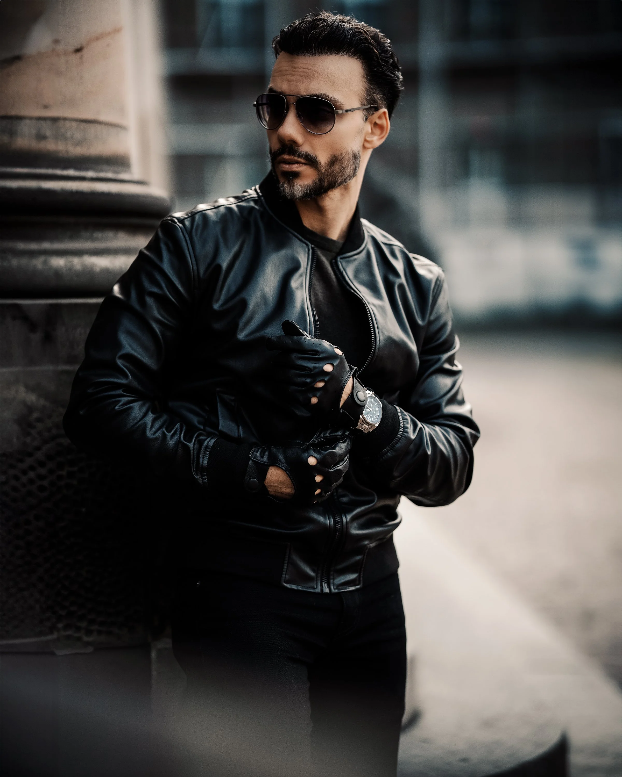 Mann mit dunklen Haaren, Bart und Sonnenbrille trägt schwarze Lederjacke in urbaner Umgebung.