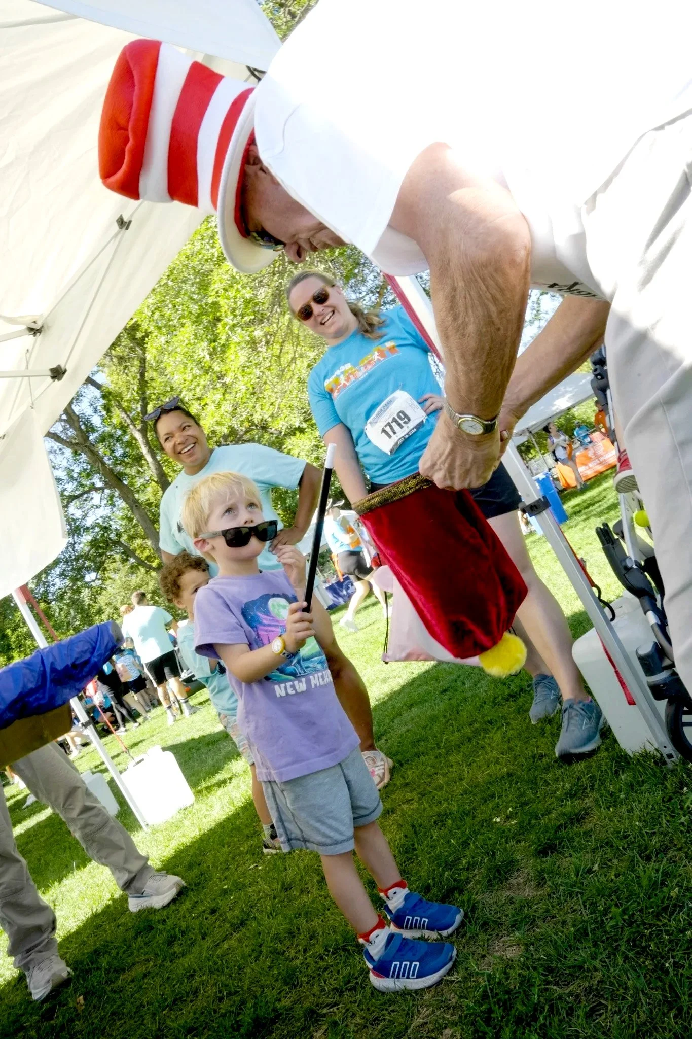 A group of children and adults at an outdoor event, with some wearing sunglasses, sunglasses, and casual clothing. One child is wearing a purple shirt, gray shorts, and blue shoes with snowman design, while an adult is dressed in white, and another adult is wearing a blue shirt. A man with a red and white hat is bending down, and others in the background are smiling and talking.