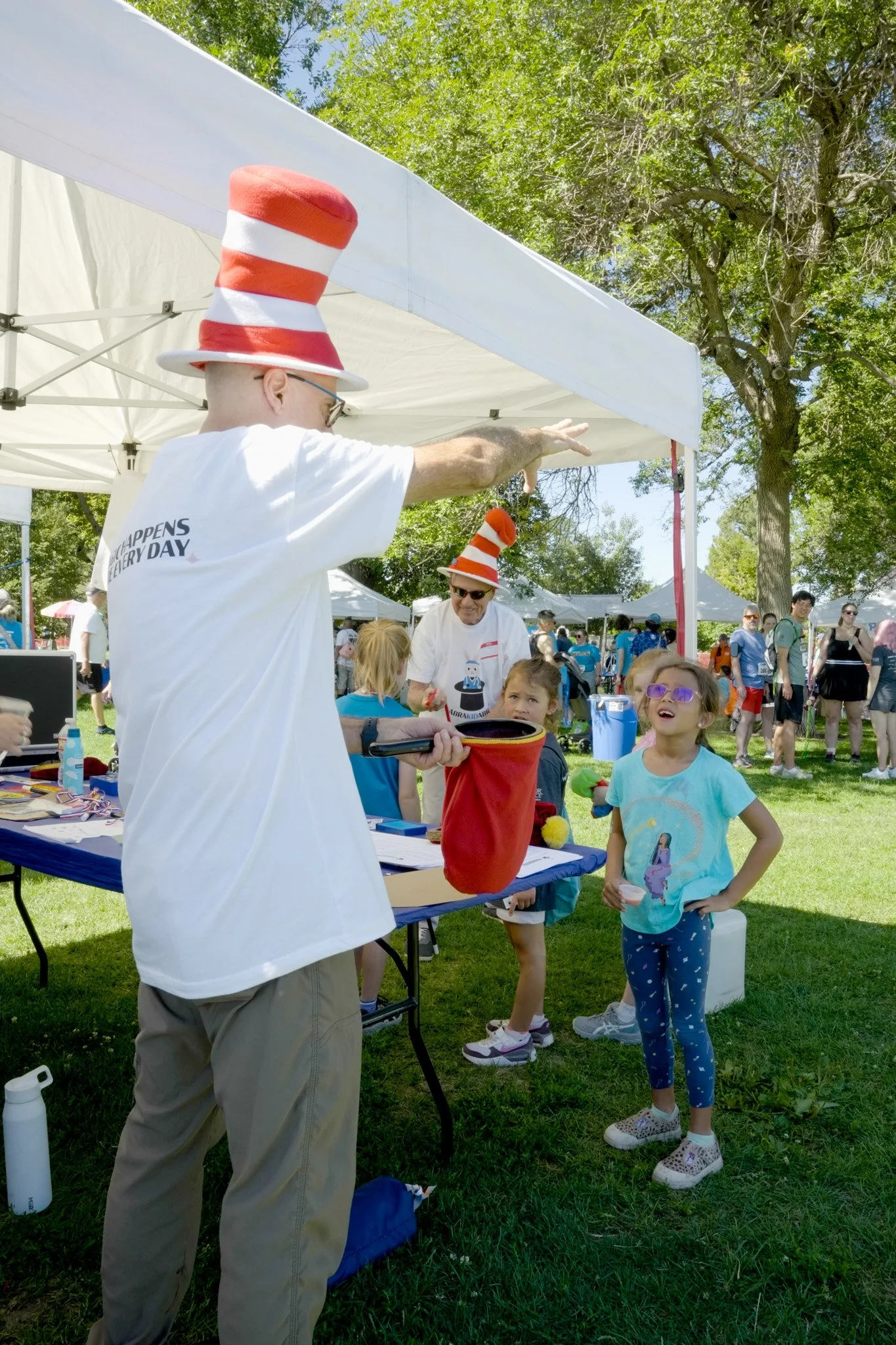 A man wearing a red and white striped Dr. Seuss hat and sunglasses pointing at a girl in a blue T-shirt with a princess on it, as other children and adults gather around at an outdoor event with tents and trees.