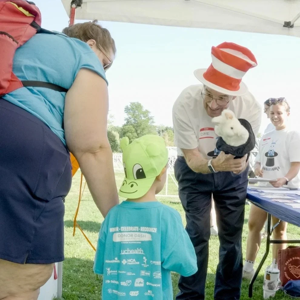 A man wearing a red and white striped hat, glasses, and a white shirt holds a white plush rabbit while talking to a child wearing a green animal hat at an outdoor event under a tent.