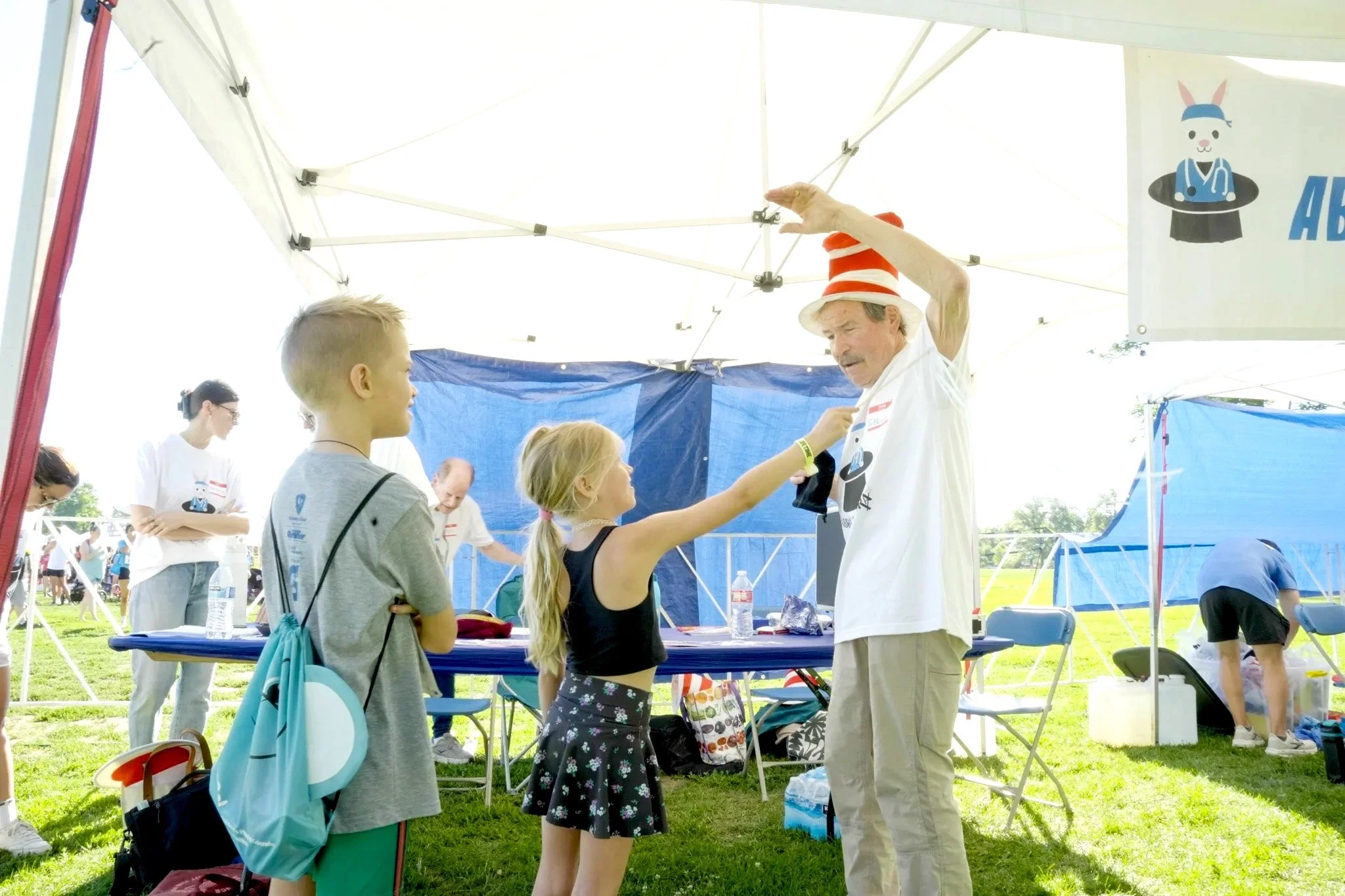 An outdoor event under a white tent with children and adults. A man wearing a Dr. Seuss-themed hat and a white T-shirt is interacting with children. Two girls are talking to him, one in a black dress and the other in patterned shorts. Other people are in the background, engaging in activities at tables and booths.