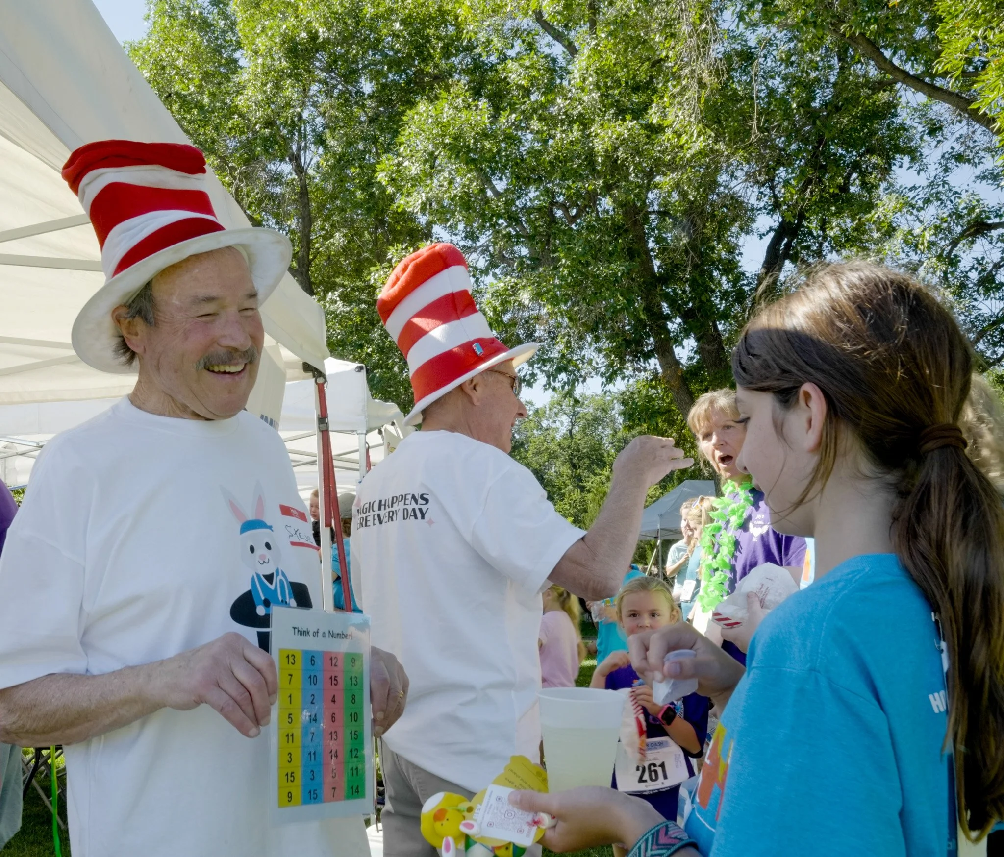 People at an outdoor event, with some wearing red and white striped hats, engaging with children and holding a colorful number chart and small stuffed animals, under green trees on a sunny day.