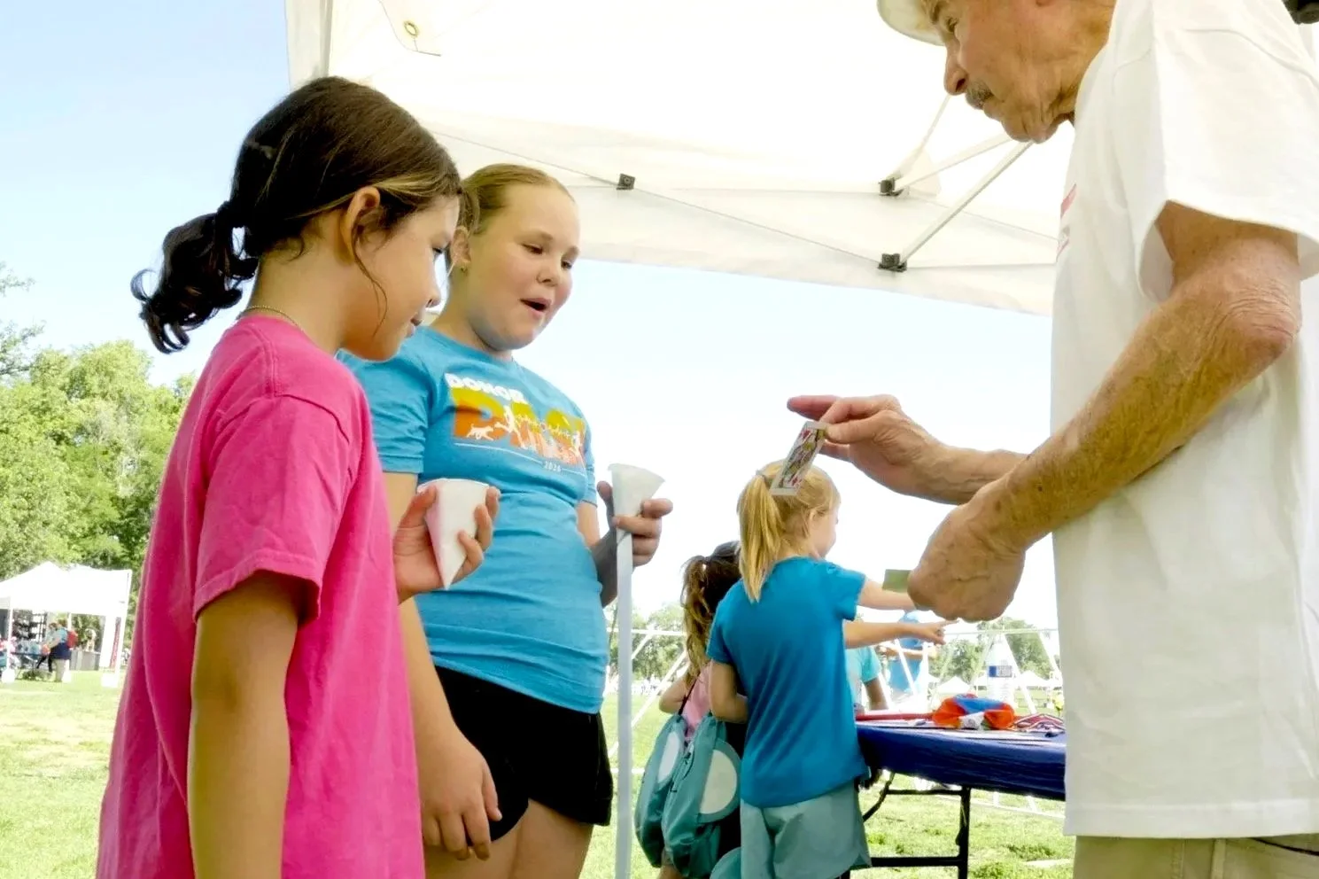 An elderly man is showing a photo to two young girls at an outdoor event under a white canopy. The girls are holding cups and looking at the photo with interest.