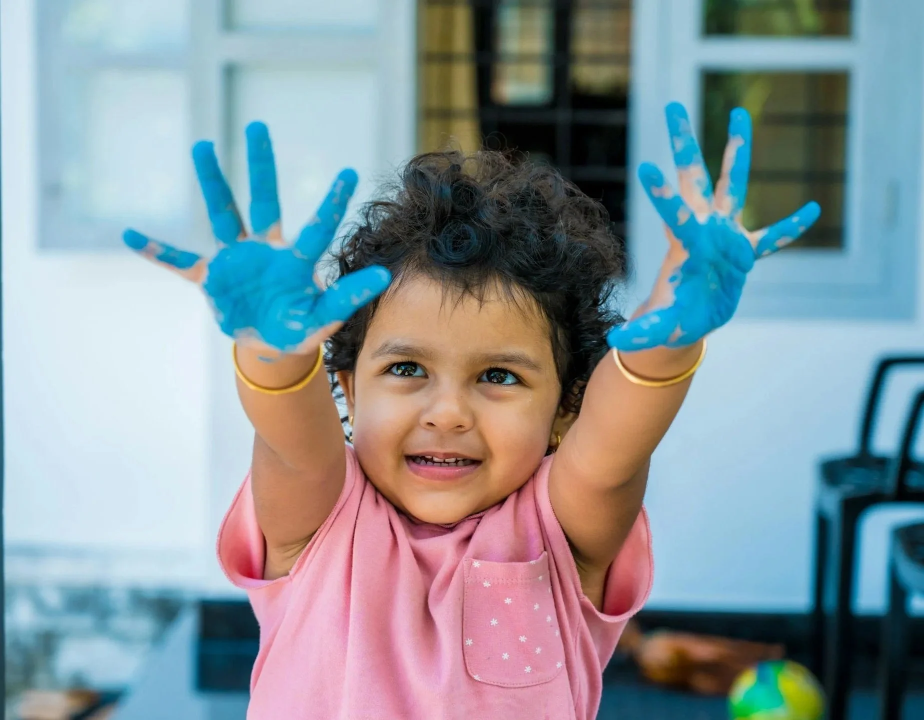 Young girl with curly hair and brown eyes, smiling and raising her hands with blue paint on them, indoors near a window and bookshelf.