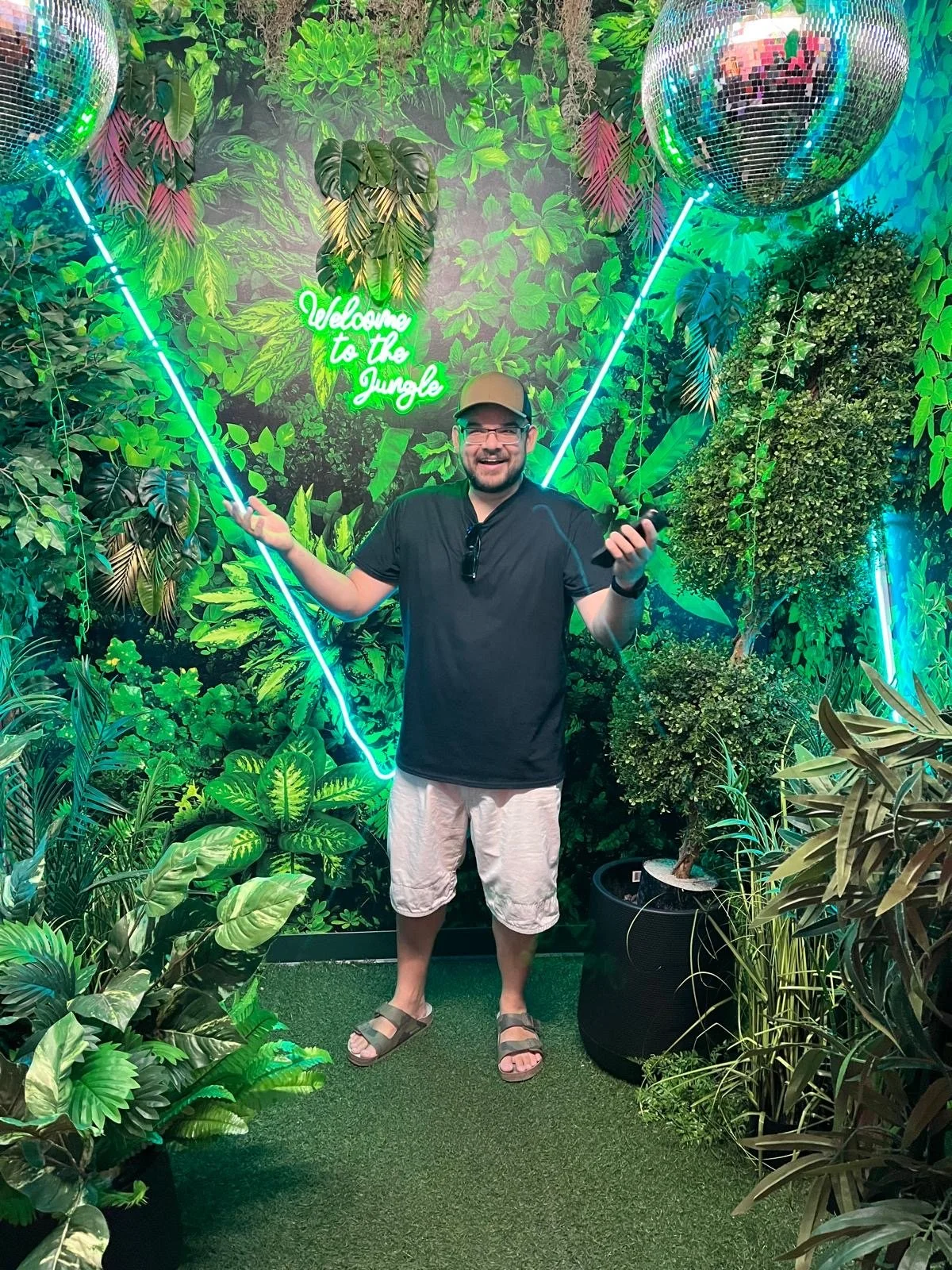 A man standing in front of a jungle-themed backdrop with green foliage, a neon sign reading 'Welcome to the Jungle,' and two mirrored disco balls. He is smiling, wearing glasses, a black t-shirt, beige shorts, sandals, and a beige hat, holding a phone in his right hand and gesturing with his left.