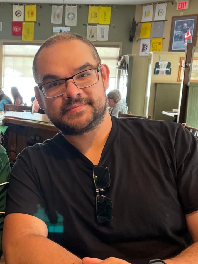 A man with glasses and a beard sitting at a restaurant table, smiling and wearing a black t-shirt, with sunglasses hanging from his collar. There are other people in the background and patriotic decorations hanging from the ceiling.