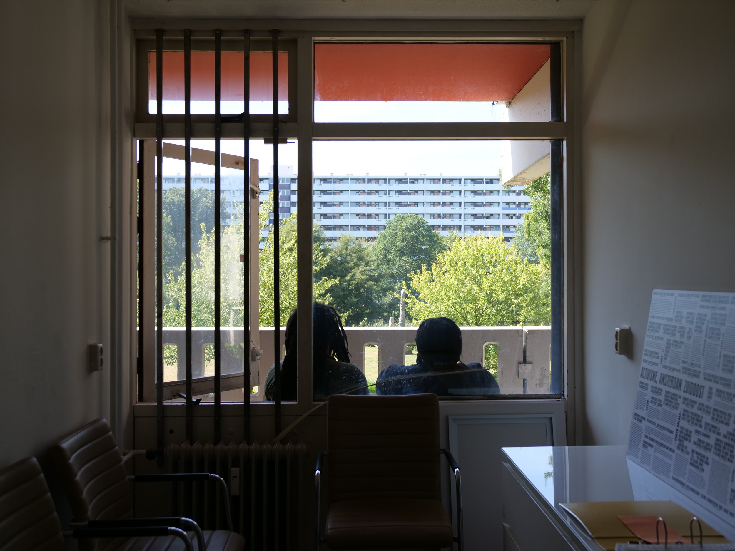 Binnenkant van de Bijlmer newsroom met een grote raam die uitkijkt op een groen landschap en de flat Gravestein, twee mensen zitten buiten op het balkon en er zitten lege stoelen binnen.