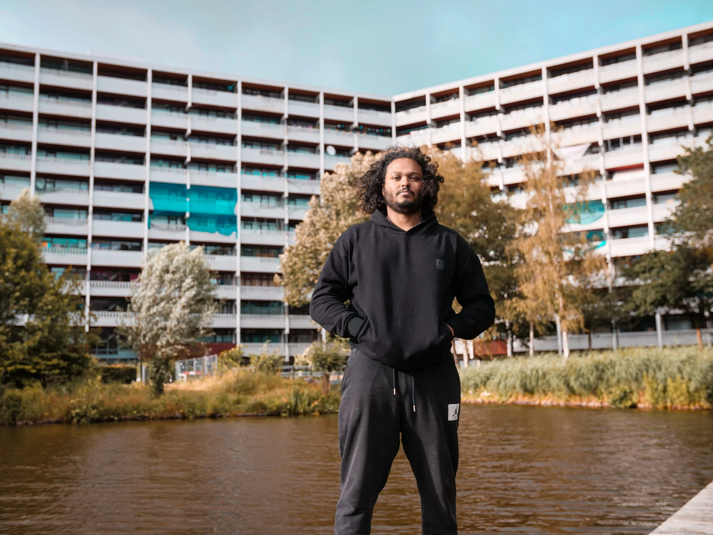 Shaquille Veldboom staat voor een rivier, met de flat Geldershoofd op de achtergrond en bomen op de voorgrond.