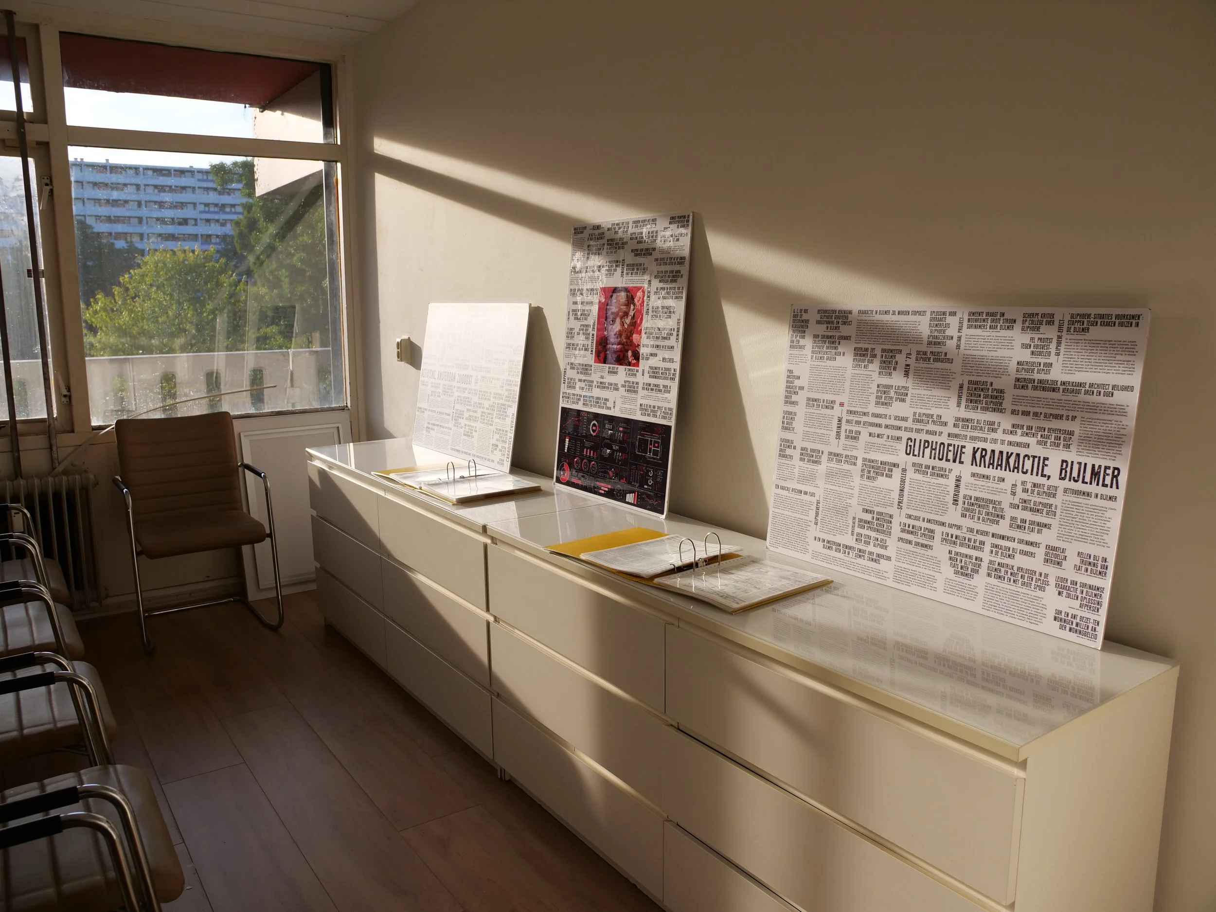 Een kamer met grote ramen die natuurlijk licht binnenlaten. Op een witte kast staan posters en documenten, waaronder een poster met een afbeelding van een persoon en tekst. Er zitten ook enkele mappen en papieren op de kast. Er staan stoelen langs de muur.