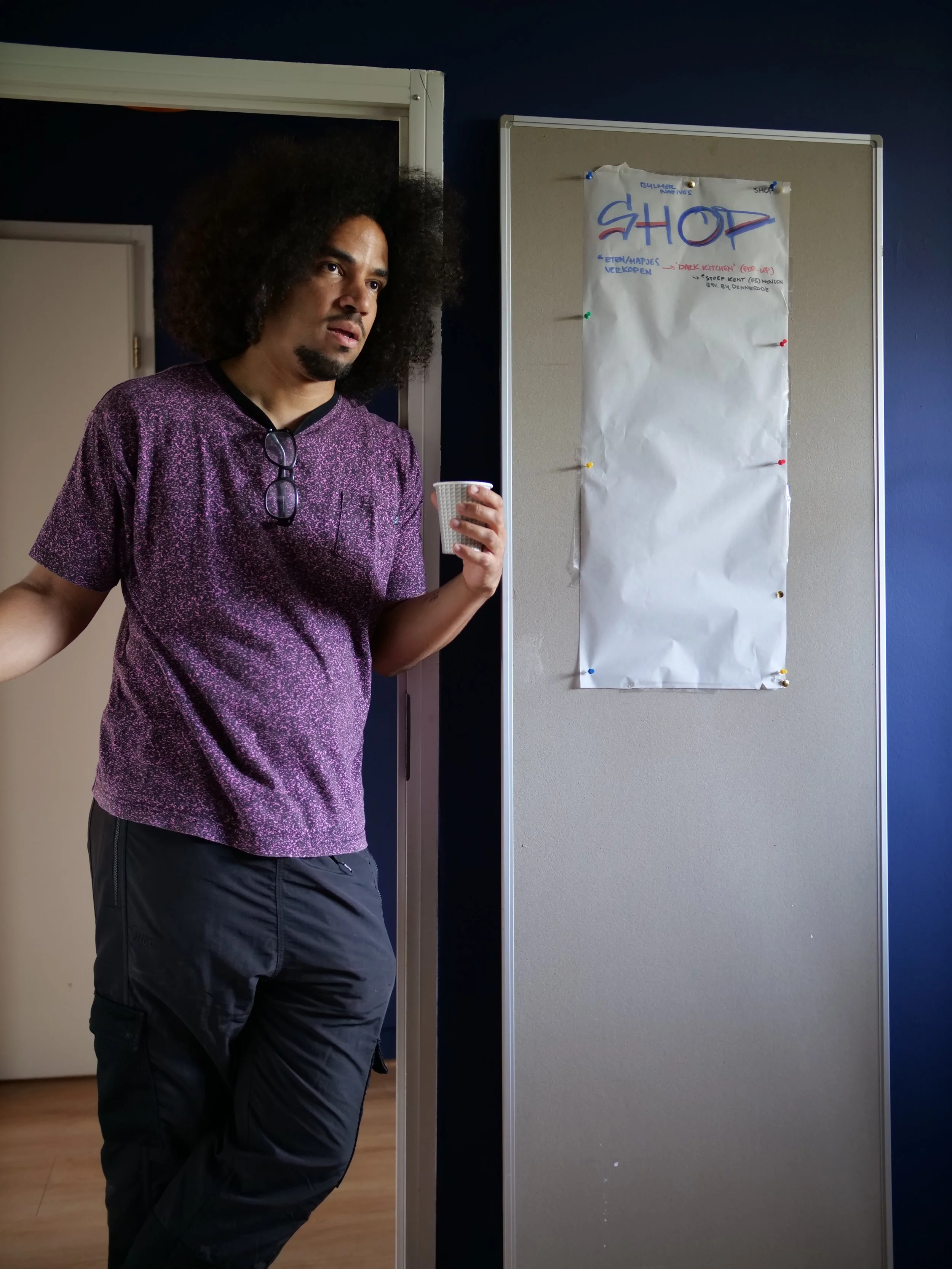 Wouter Pocornie met krullend haar die in een kamer staat en een papieren beker vasthoudt naast een blanco instructiebord op de muur.