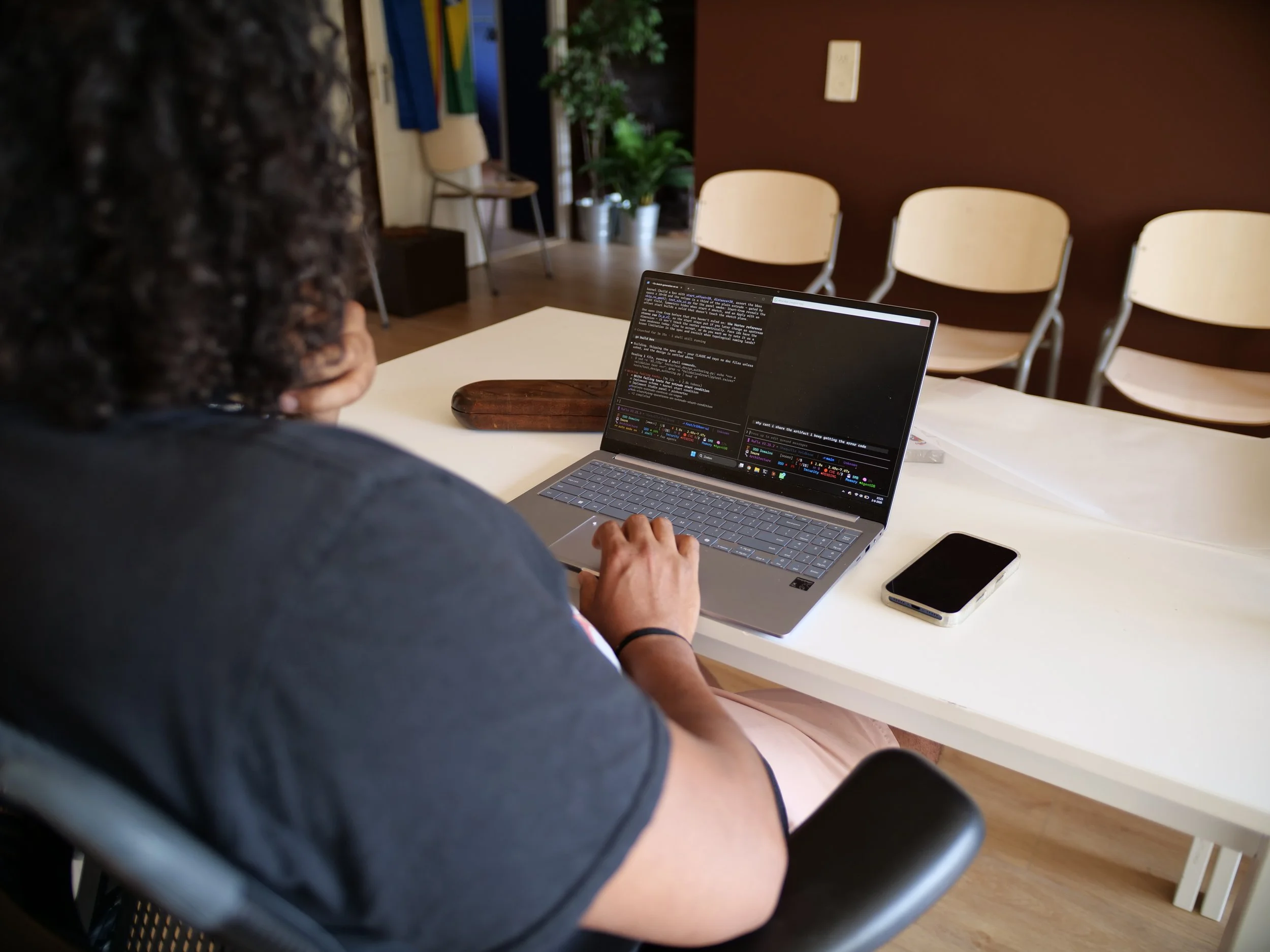 Shaquille Veldboom zit aan een witte tafel met een laptop open, die tekst en code toont. Op de tafel liggen een smartphone en een houten bureaublad. De omgeving lijkt een informele kantoorruimte of vergaderruimte met witte stoelen en planten.