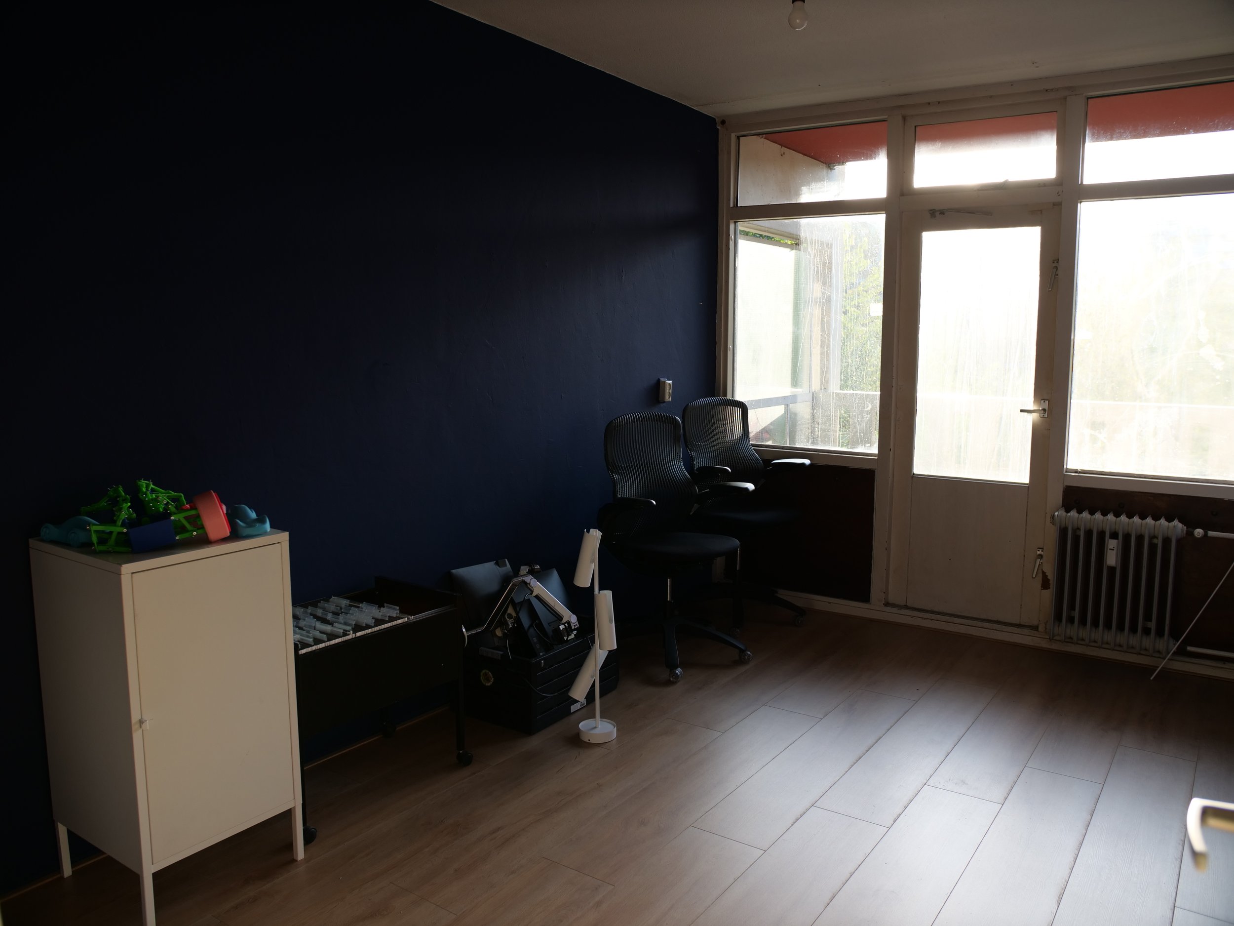 Een lege kamer met een grote raampartij en een donkerblauwe muur, enkele zwarte stoelen, en een witte kast met kinderfietsjes en 3D-geprint speelgoed.