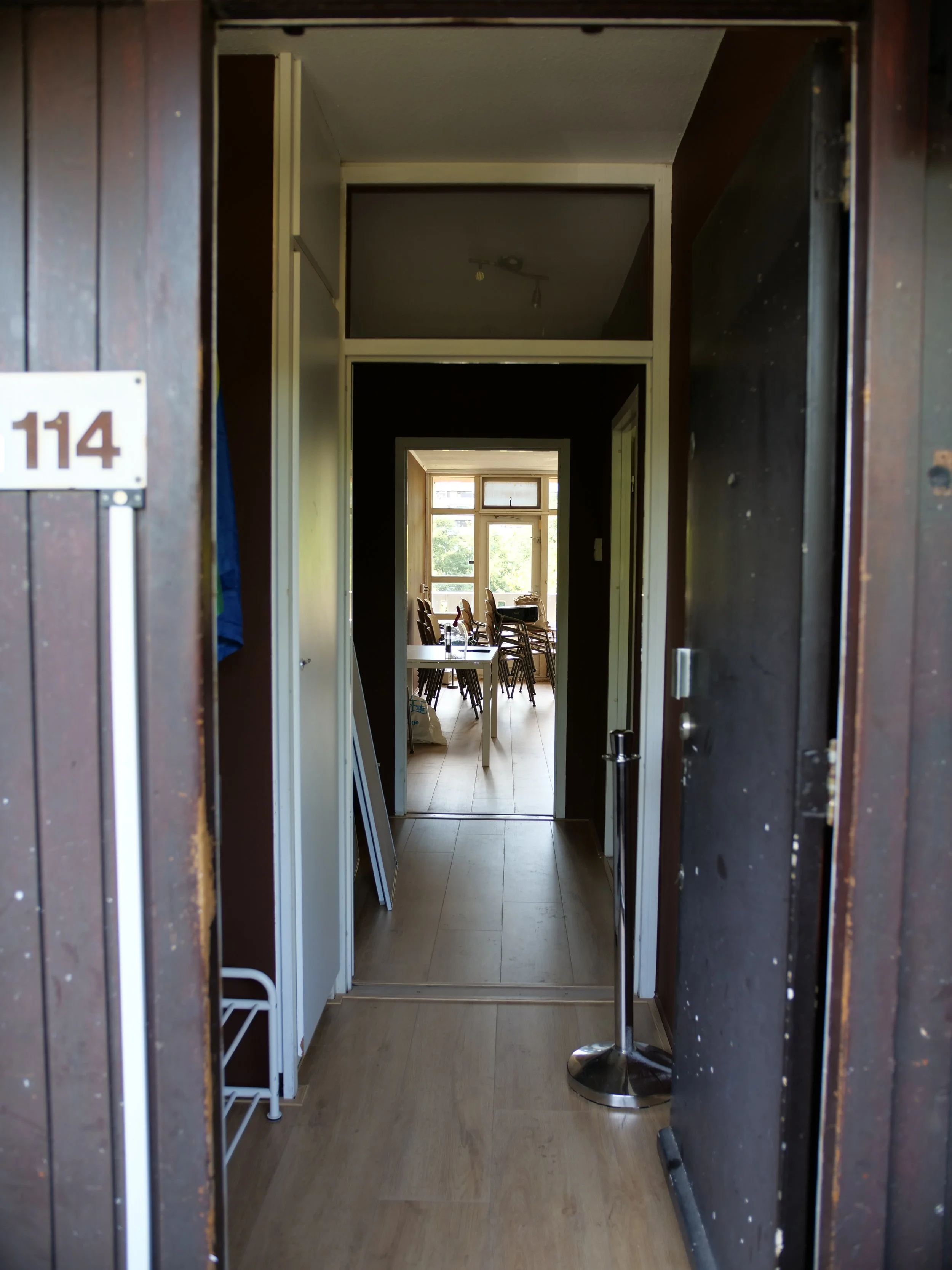 De ingang met een lange gang. In de woonkamer staan enkele stoelen rondom een tafel, grote ramen waardoor natuurlijk licht binnenkomt.