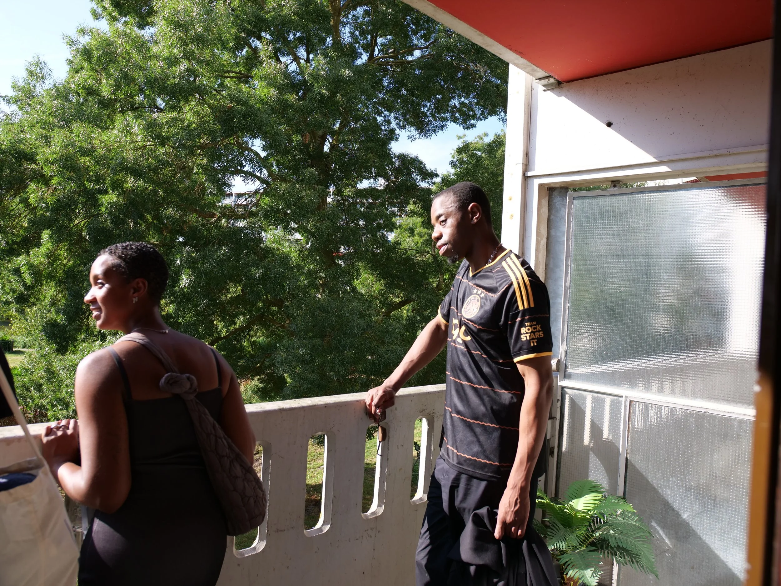 Twee bezoekers staan op het balkon met groene bomen op de achtergrond. Eén persoon kijkt naar beneden en de andere, met een zwart en geel shirt, kijkt in de verte.