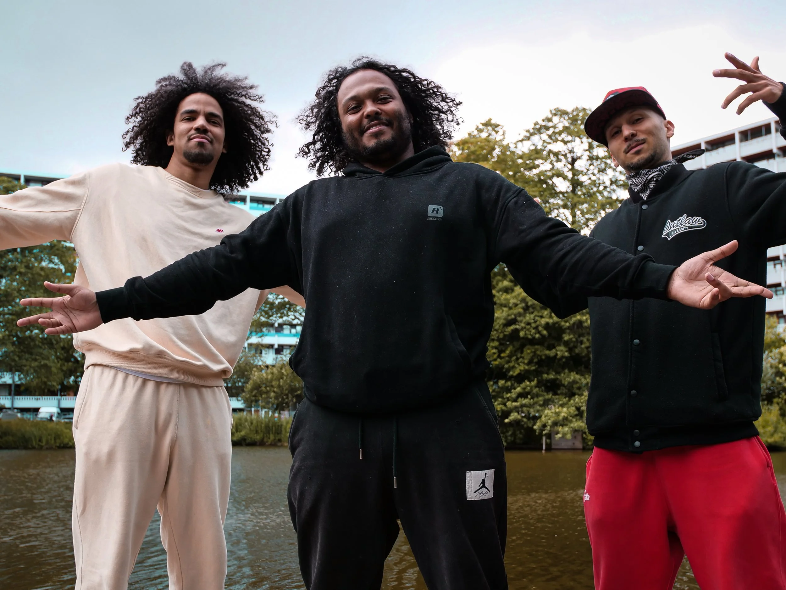 Wouter Pocornie, Shaquille Veldboom en Karim KHamis staan samen buiten voor een rivier, met een park en de flats Gravestein en Geldershoofd op de achtergrond, terwijl ze vriendelijk naar de camera lachen.