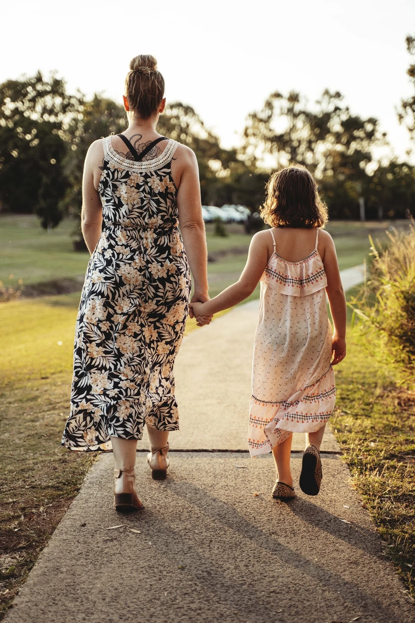 A woman and a young girl walk hand in hand on a sidewalk in a park during sunset, surrounded by greenery and trees.