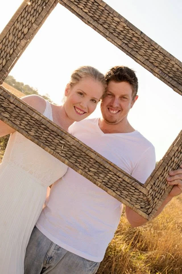 A smiling young woman and man standing outdoors in a field, holding a large empty picture frame around their faces during sunset.