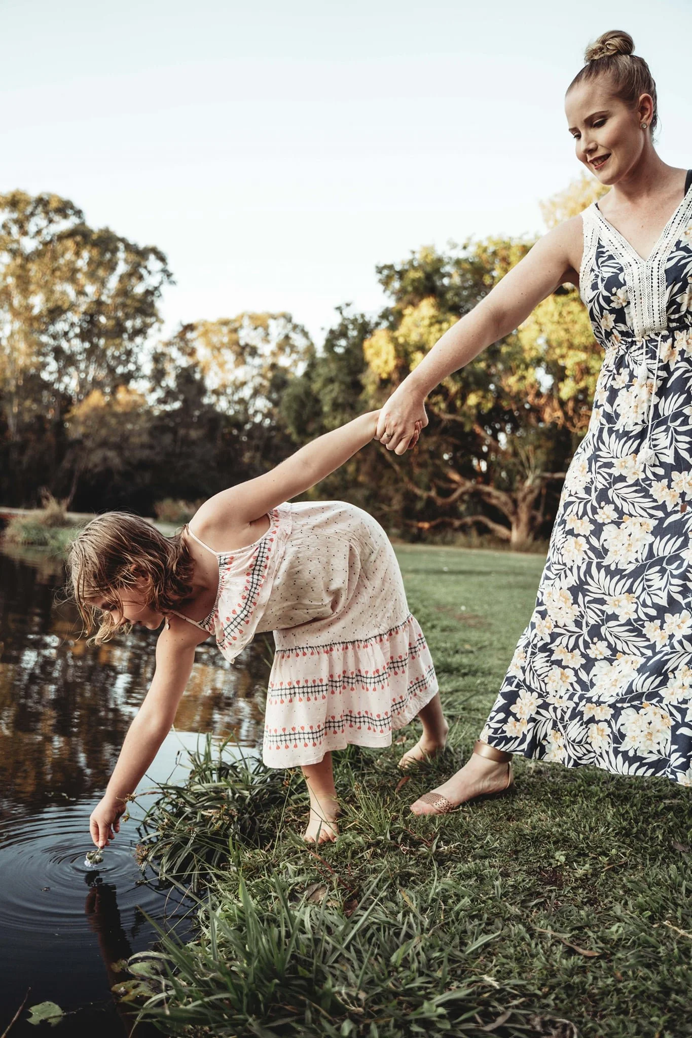 A woman and a young girl by a pond, with the woman holding the girl's hand as they lean over the water, engaging in an outdoor activity during sunset.
