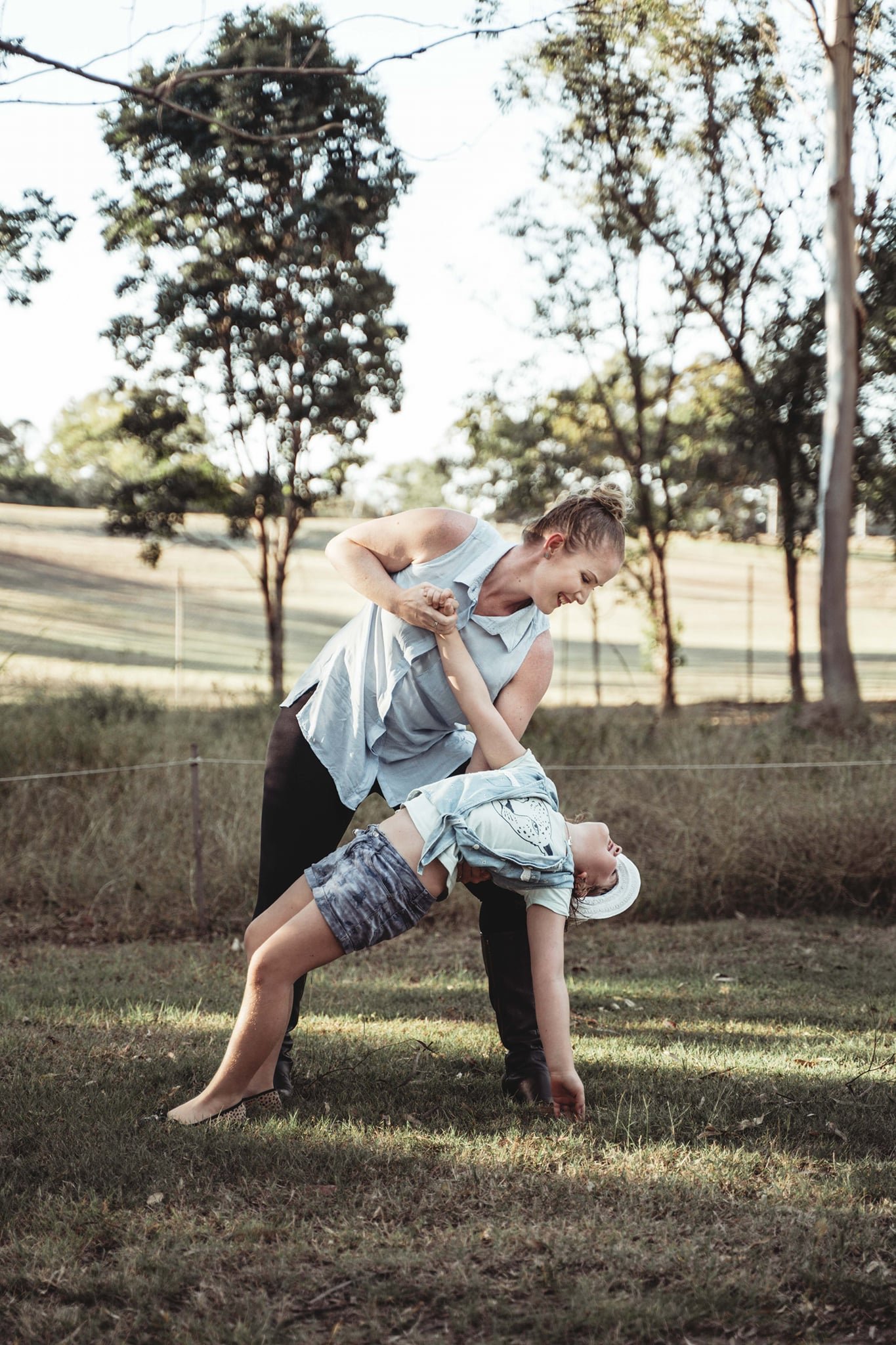 A woman holding a young boy in a yoga pose outdoors in a park with trees in the background.