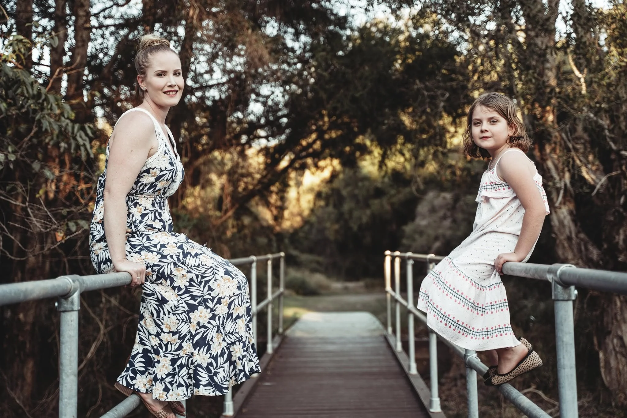 A woman and a young girl sitting on a metal railing on a bridge in a wooded area during sunset.