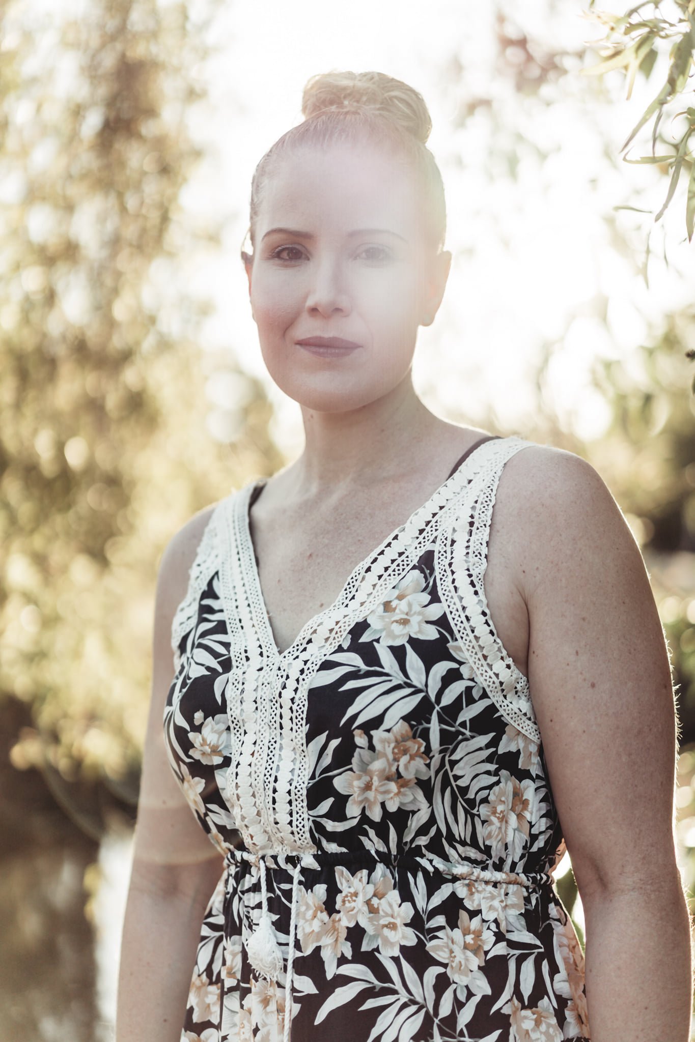 A woman with blonde hair in a high bun, wearing a black and white dress with floral and leaf patterns, standing outdoors with trees and sunlight in the background.