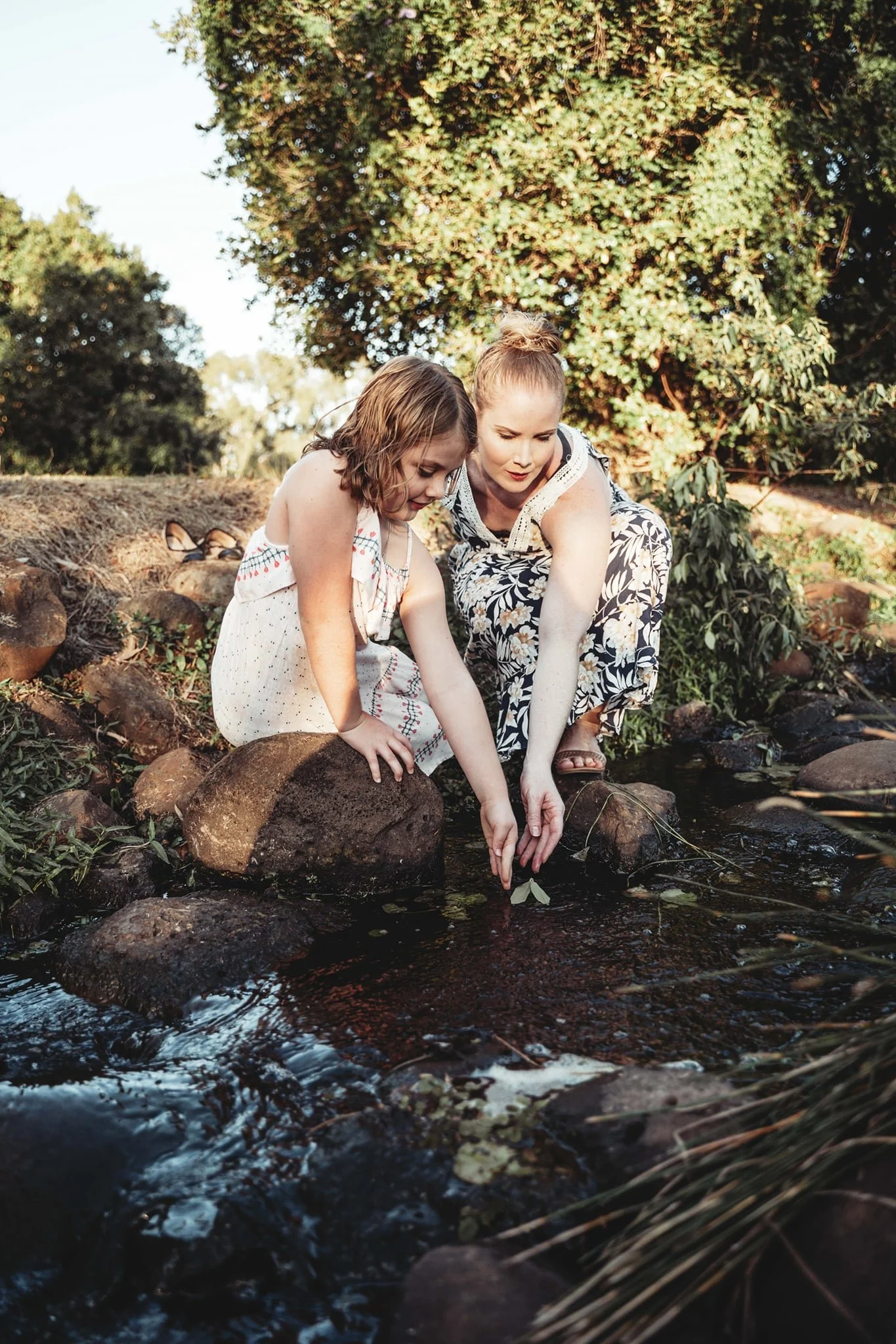 A woman and a young girl are kneeling by a creek, pointing at the water together, surrounded by rocks and trees in a natural outdoor setting.