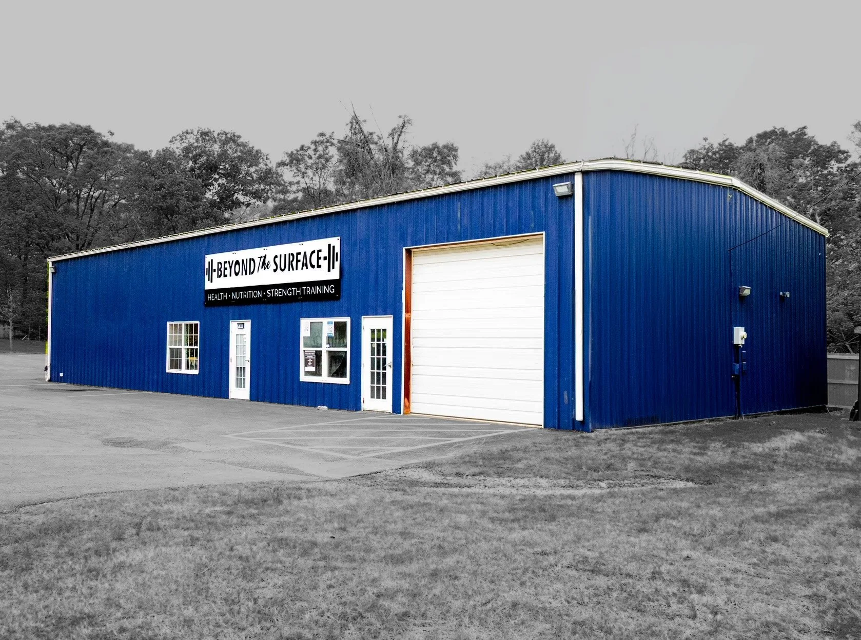 Blue industrial building with a sign that reads 'Beyond the Surface' and lists health, nutrition, and strength training, with a white garage door, several windows, and a parking area in the foreground. The background includes trees and a gray sky.