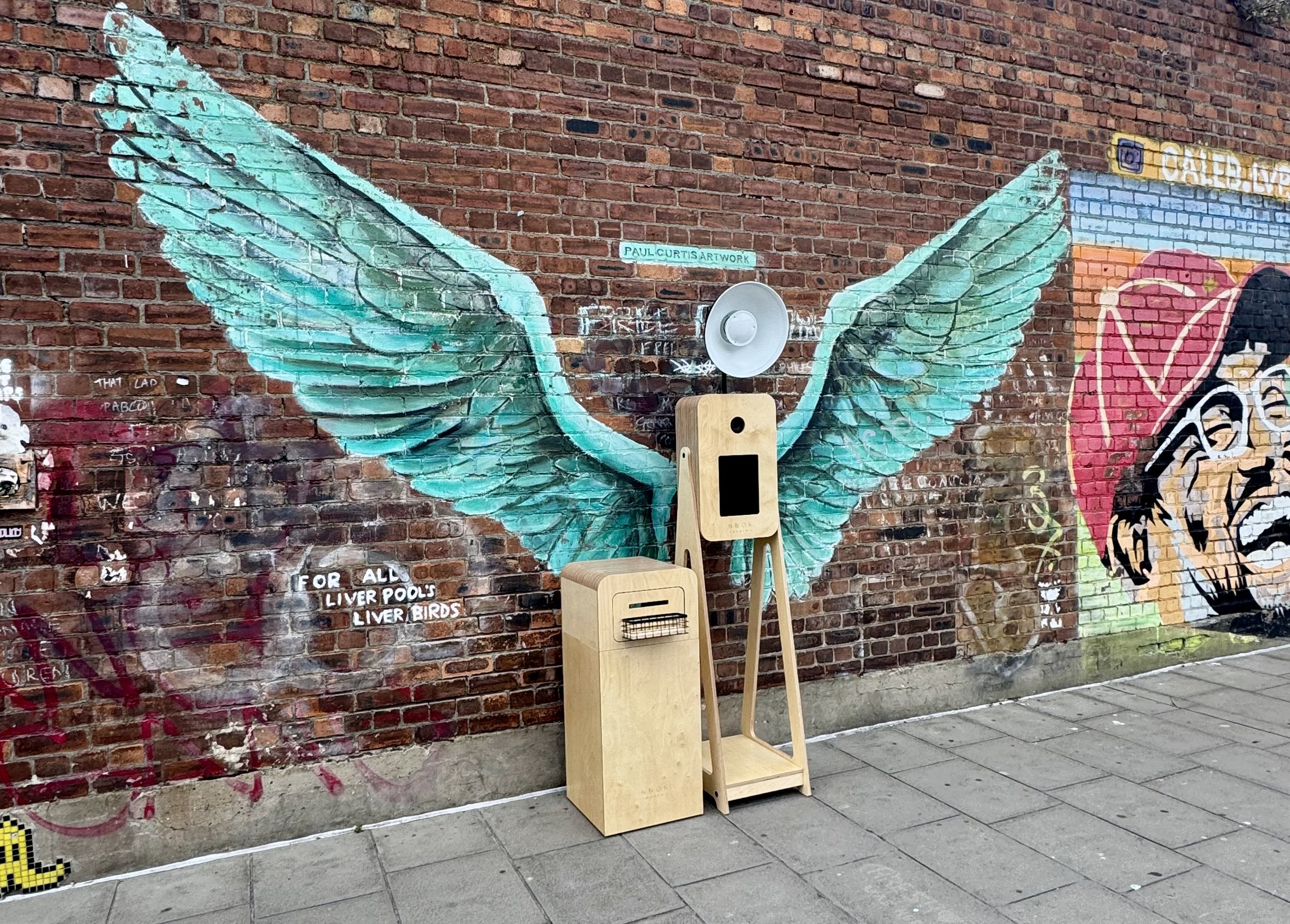 Graffiti mural of teal-colored angel wings painted on a brick wall, with a sign that reads 'PAUL CURTIS ARTWORK' in the center. There are two wooden chairs and a megaphone in front of the mural. Part of other graffiti artwork is visible on the right.