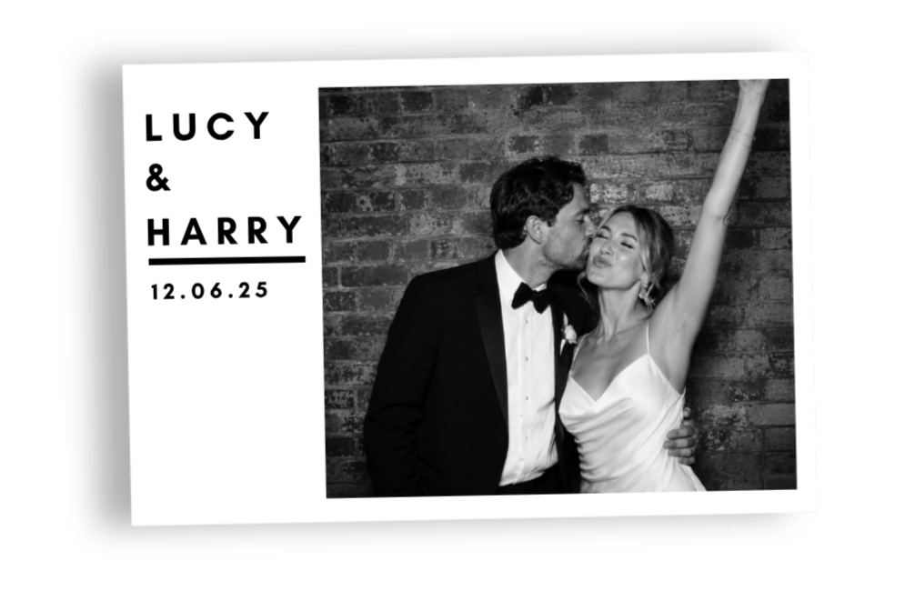 Black and white wedding photo of a couple, Lucy and Harry, with Lucy raising her arm and Harry kissing her cheek, against a brick wall background.