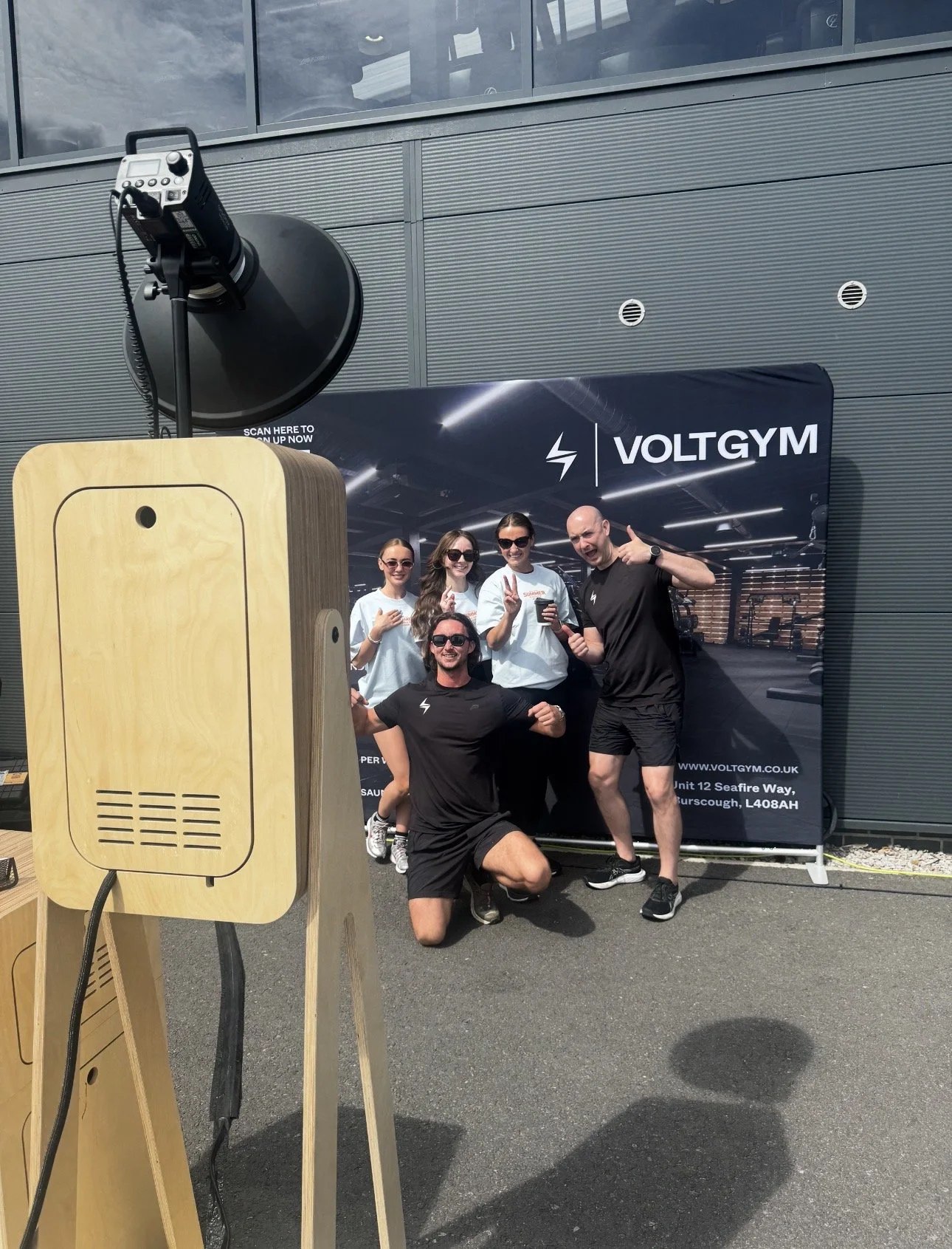 A group of five people, three women and two men, posing outdoors in front of a VoltGym banner. They are smiling, some making peace signs, one man is kneeling, and they are dressed in black and white workout clothes. Taken by Nook booth Photo Booth.