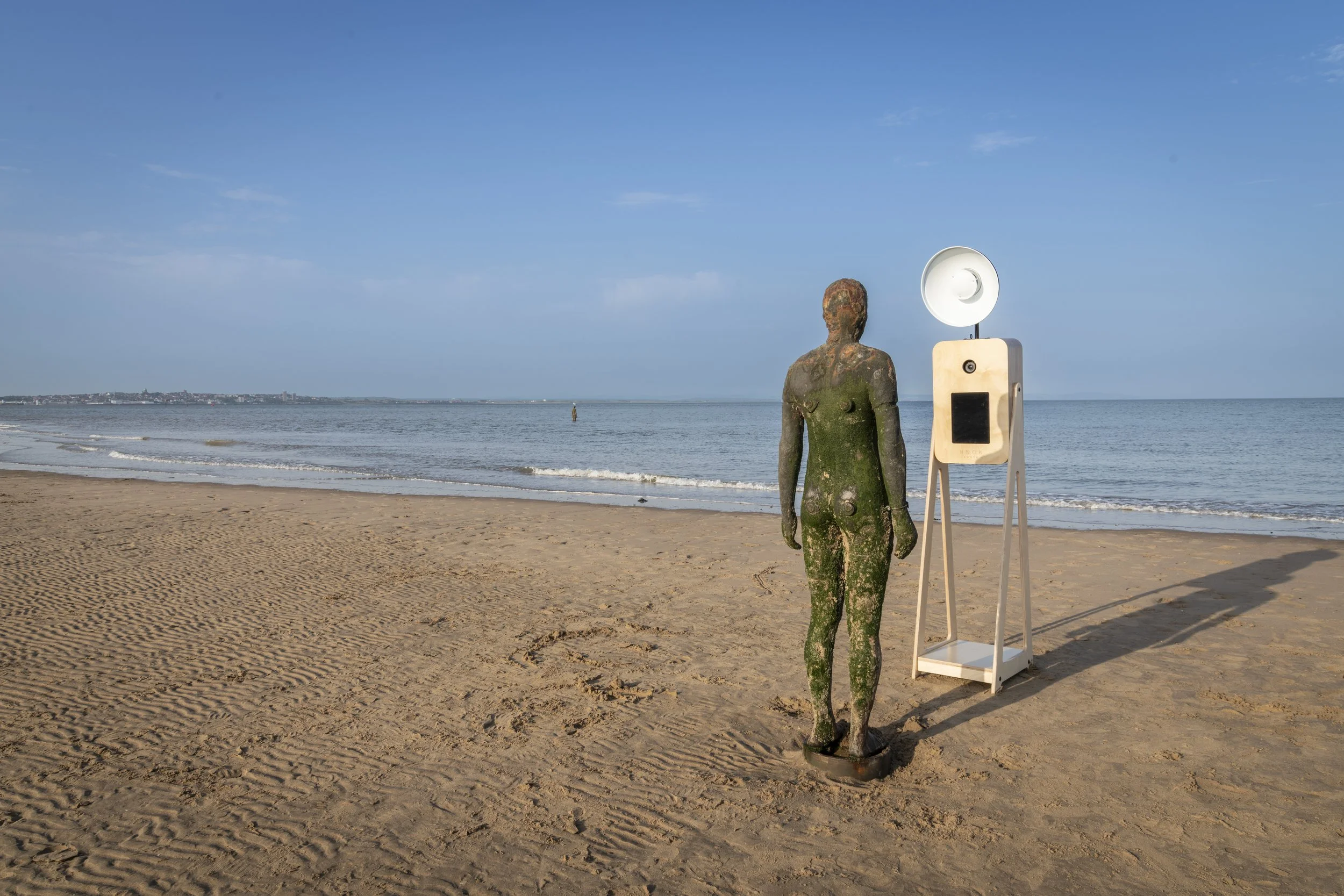 A sculpture of a human figure and a Nook Booth on a beach, with water and a distant shoreline under a blue sky.