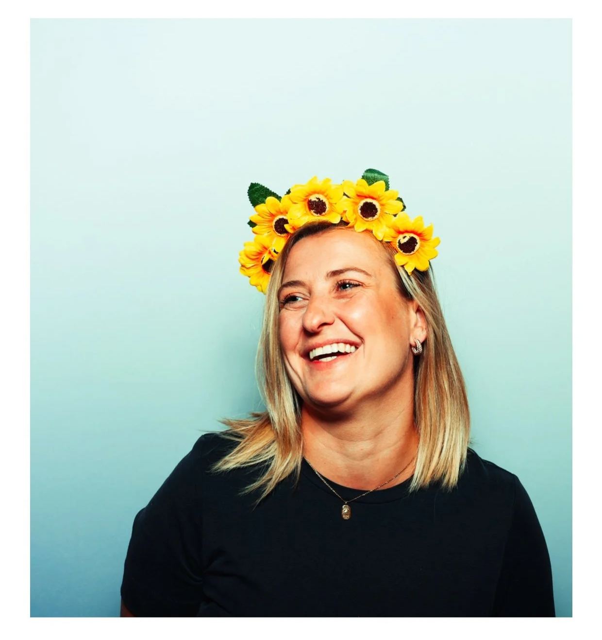 A woman with blonde hair wearing a black shirt and a sunflower crown, smiling against a light blue background. Taken by Nook booth Photo Booth