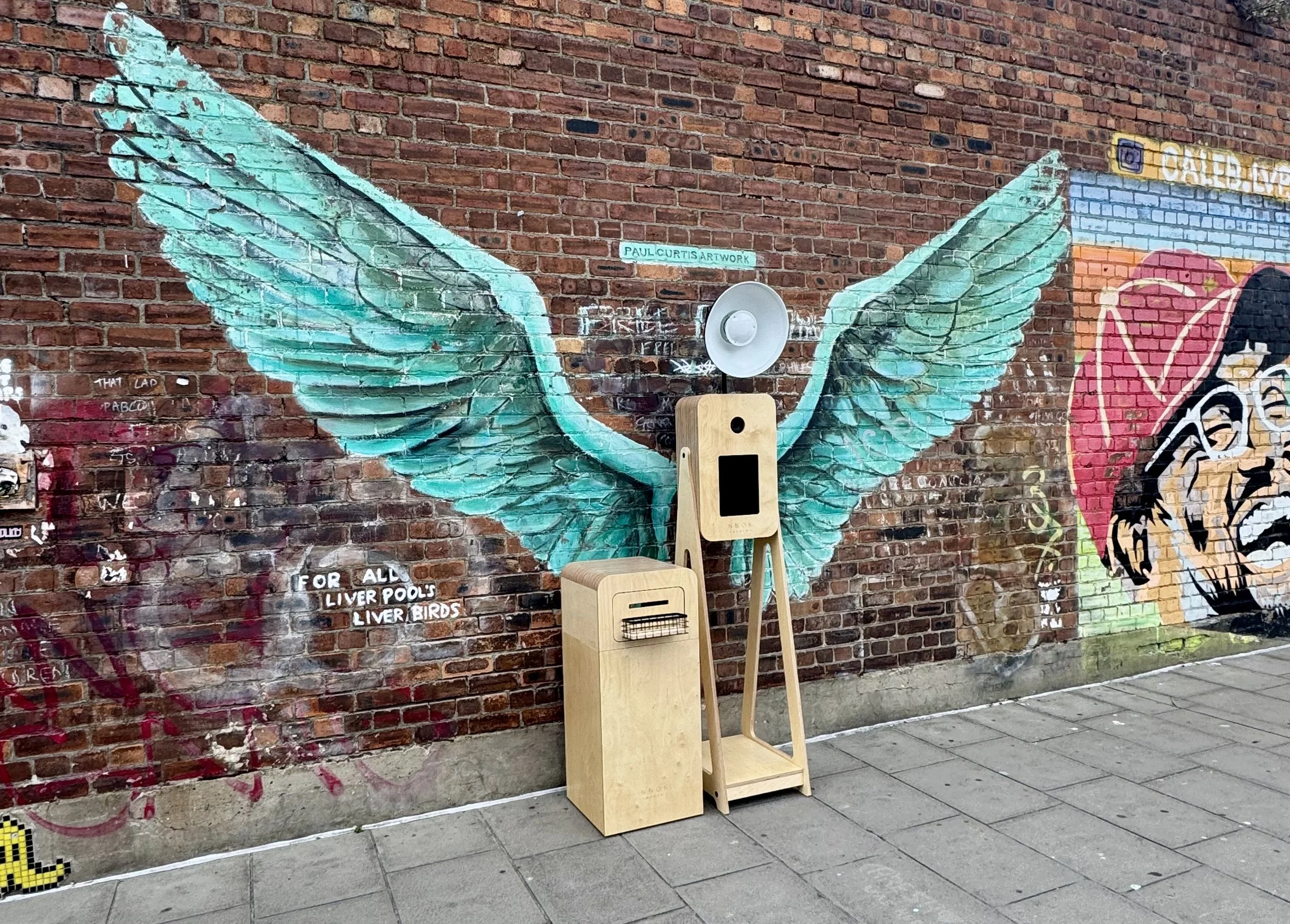 Nook Booth Photo Booth in front of  mural of teal-colored angel wings painted on a brick wall, Baltic Triangle, Liverpool.