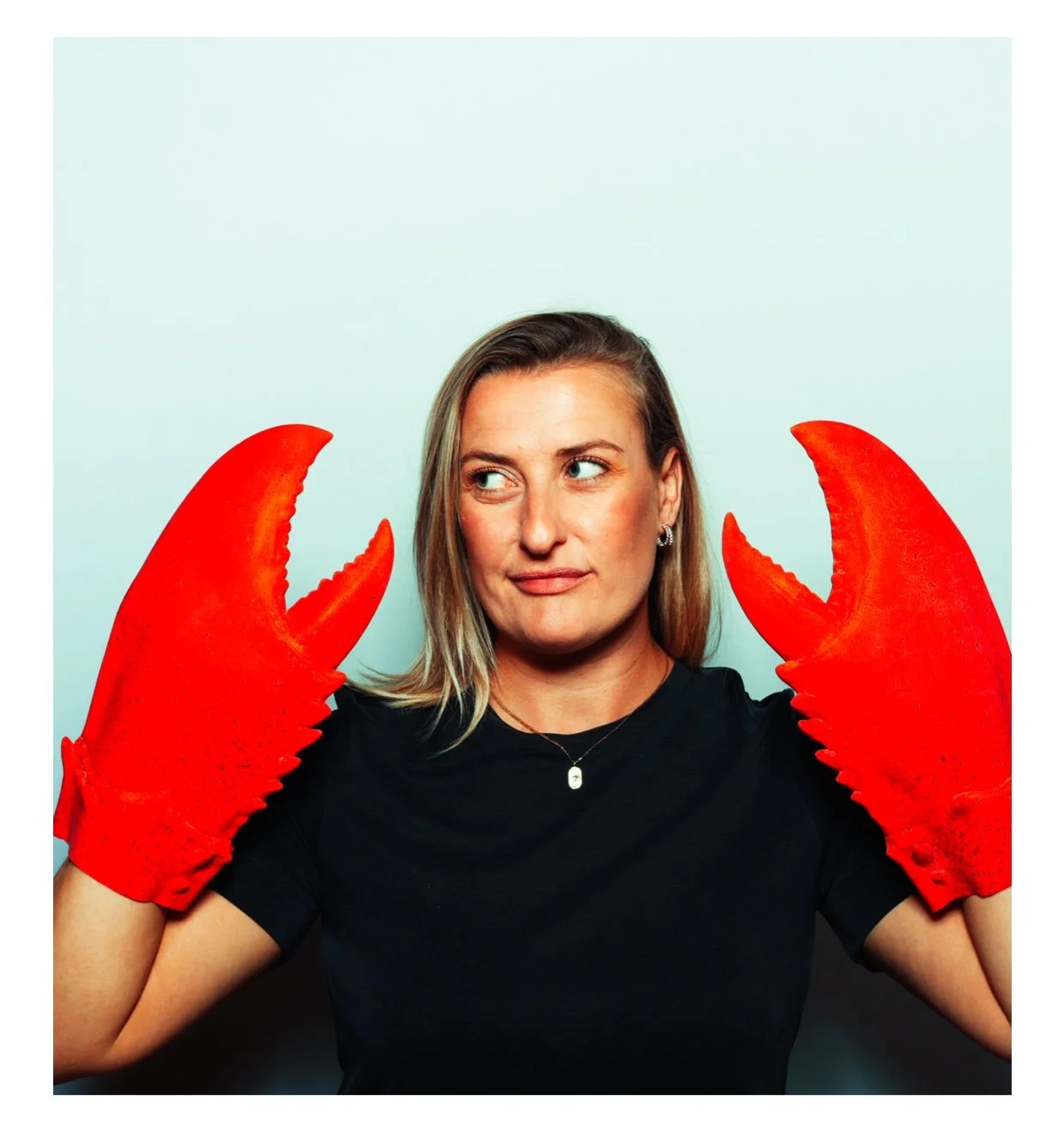 A woman with blonde hair wearing a black shirt and red lobster gloves, holding her hands up near her head, looking sideways with a playful expression, against a plain white background. Taken by Nook booth Photo Booth
