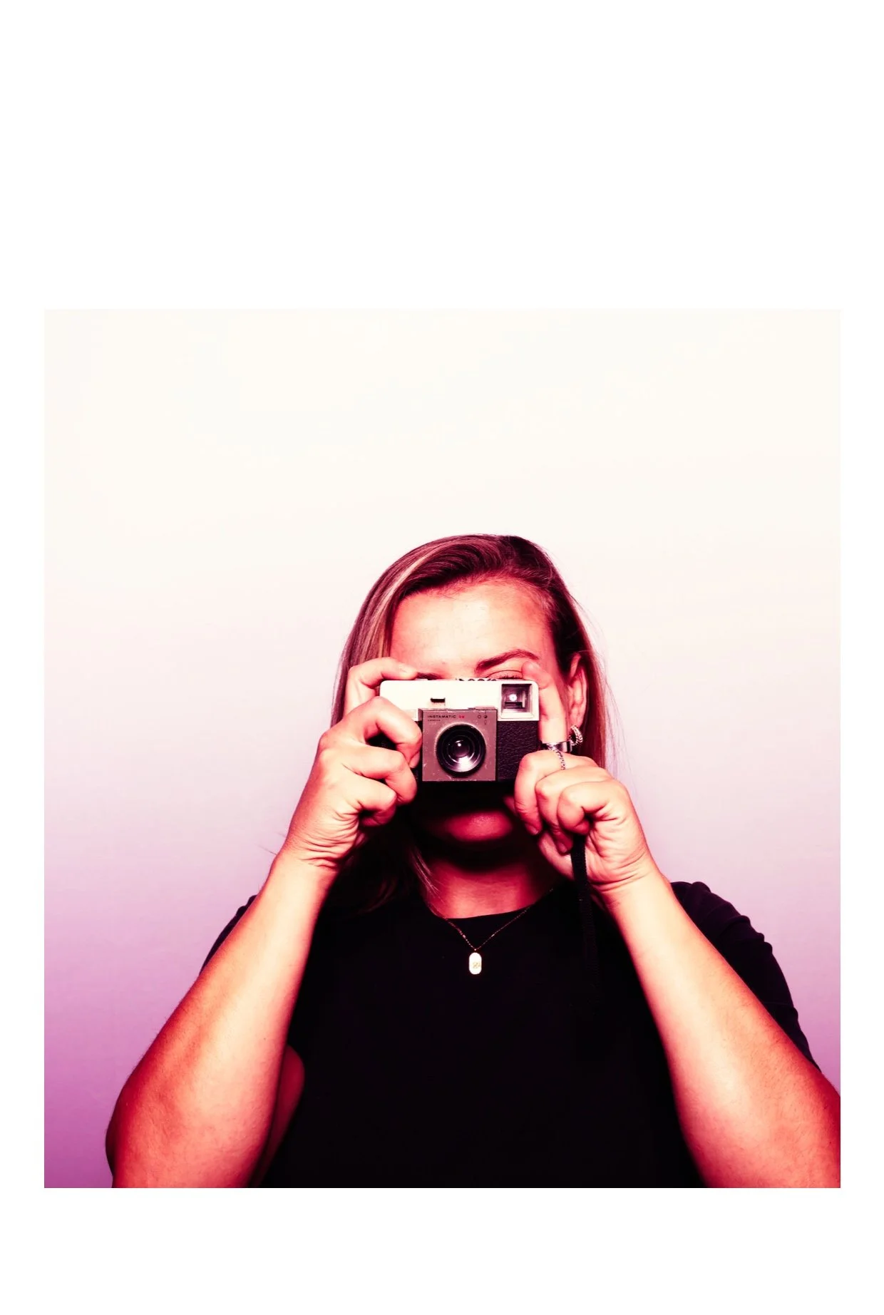 Woman with long hair taking a photo using a vintage camera, wearing a black shirt, jewelry, and standing against a plain background. Taken by Nook booth Photo Booth.