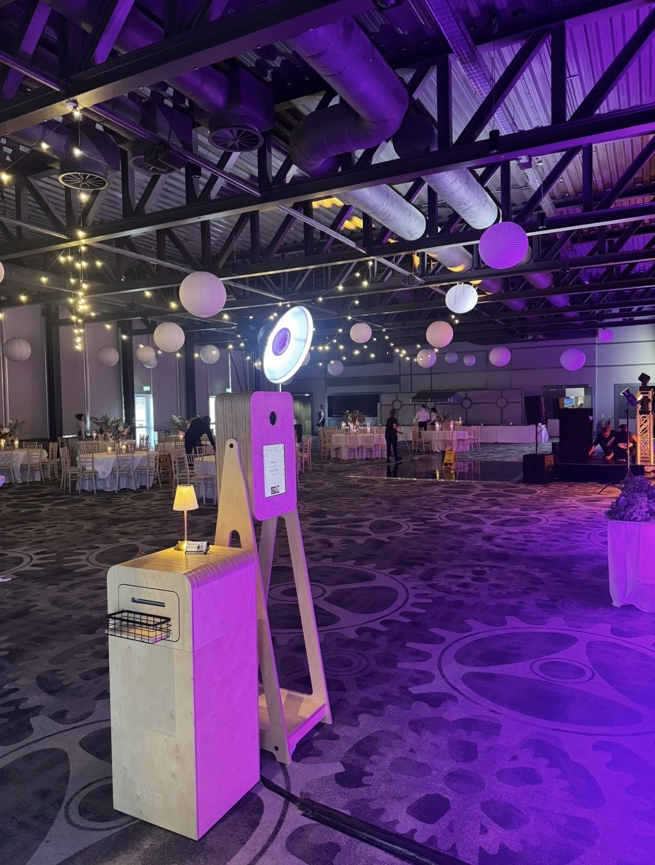 A decorated indoor event space with purple and white lighting, tables set for dining, paper lanterns hanging from the ceiling, and people preparing the venue.titanic hotel Photo Booth hire