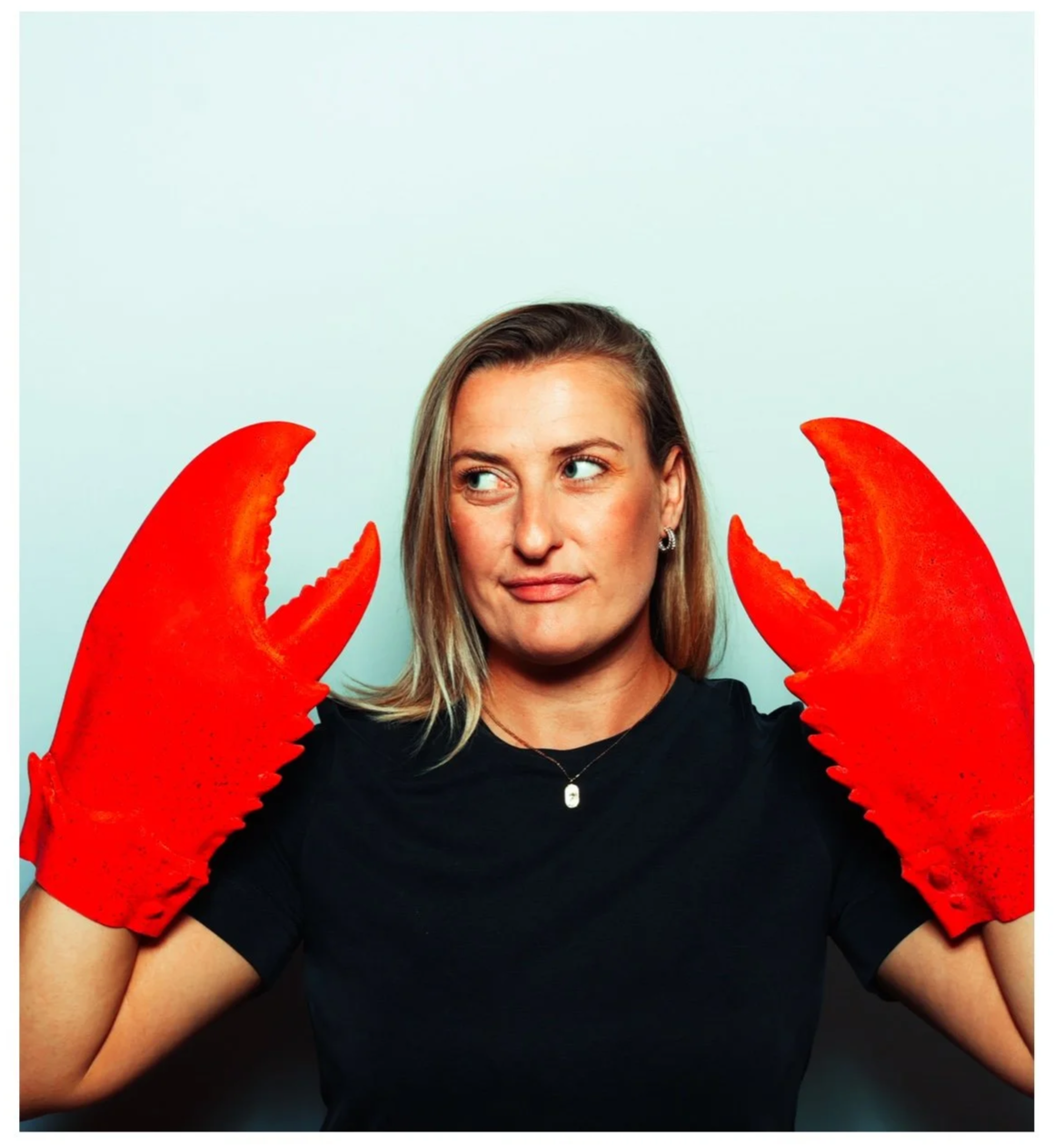 A woman with shoulder-length blonde hair wearing a black shirt and red lobster claw gloves, looking to her left with a neutral expression, against a plain light background. Taken by Nook booth Photo Booth