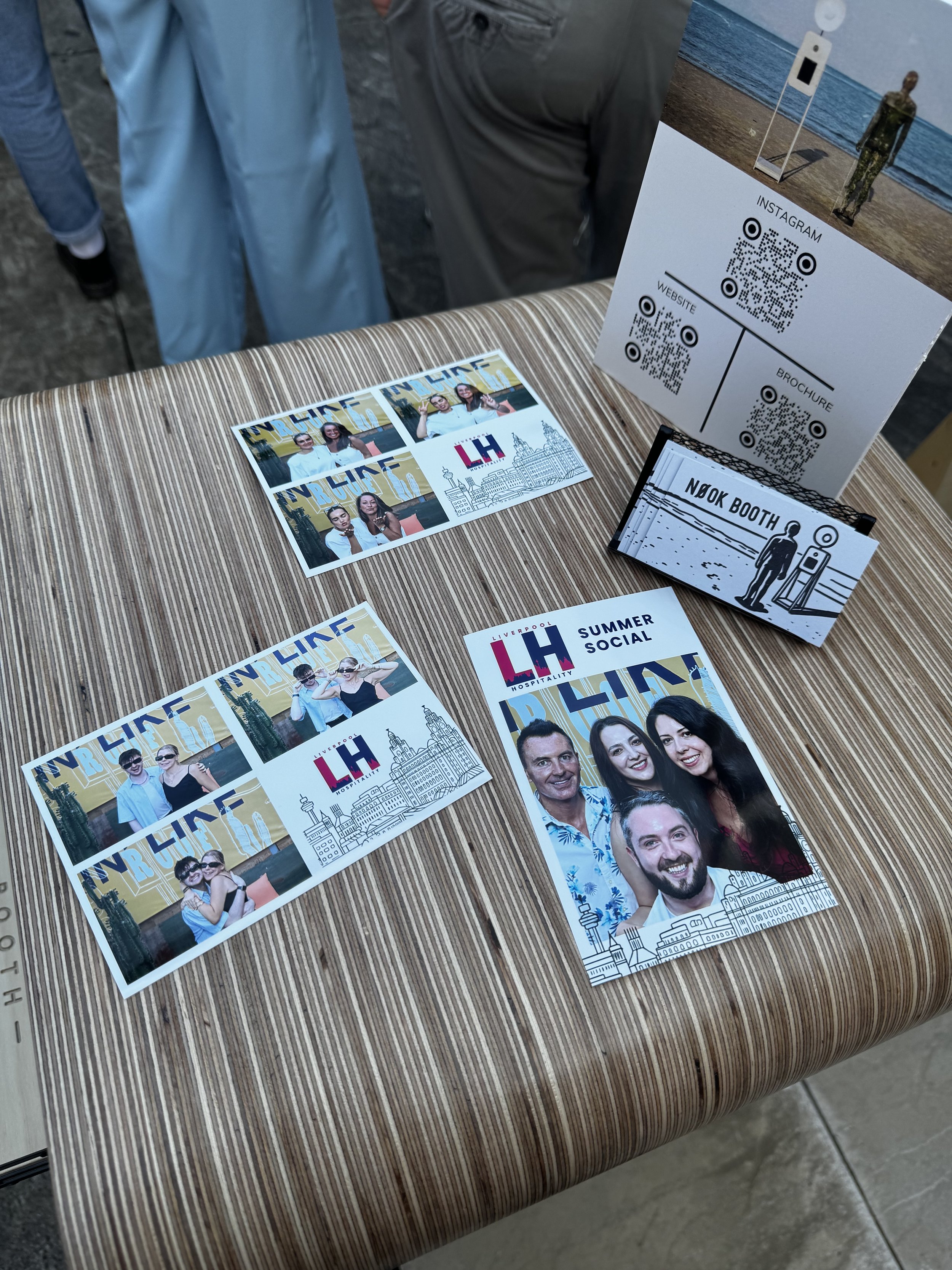 Photo of lip balm samples, flyers, and promotional material on a striped table, with a stand-up display featuring QR codes, a brochure, and a photograph of a person at the beach in the background. Taken by Nook booth Photo Booth.