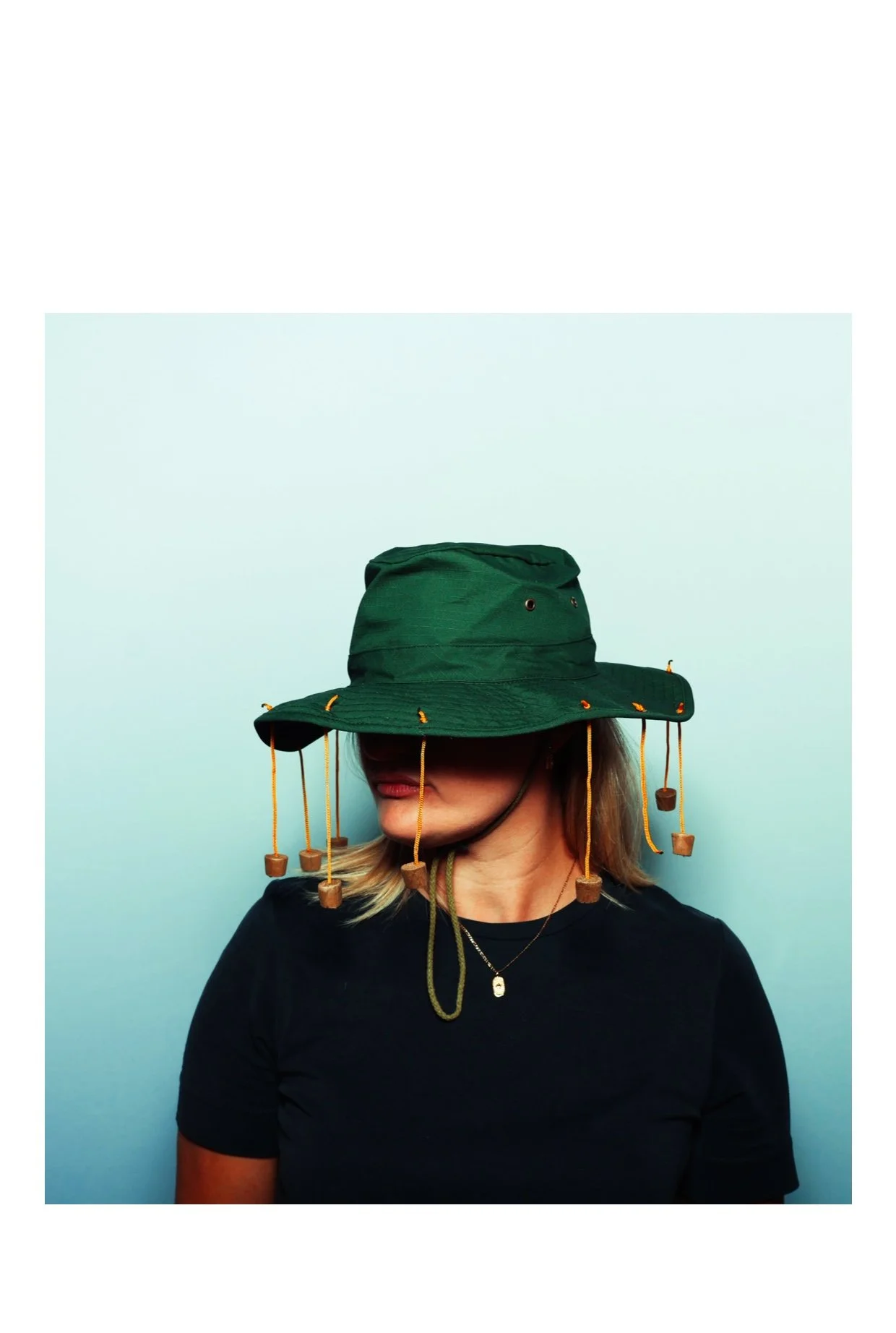 Woman wearing a green outdoor hat with yellow drawstrings and small hanging wooden pieces, facing left against a light blue background. Taken by Nook booth Photo Booth.