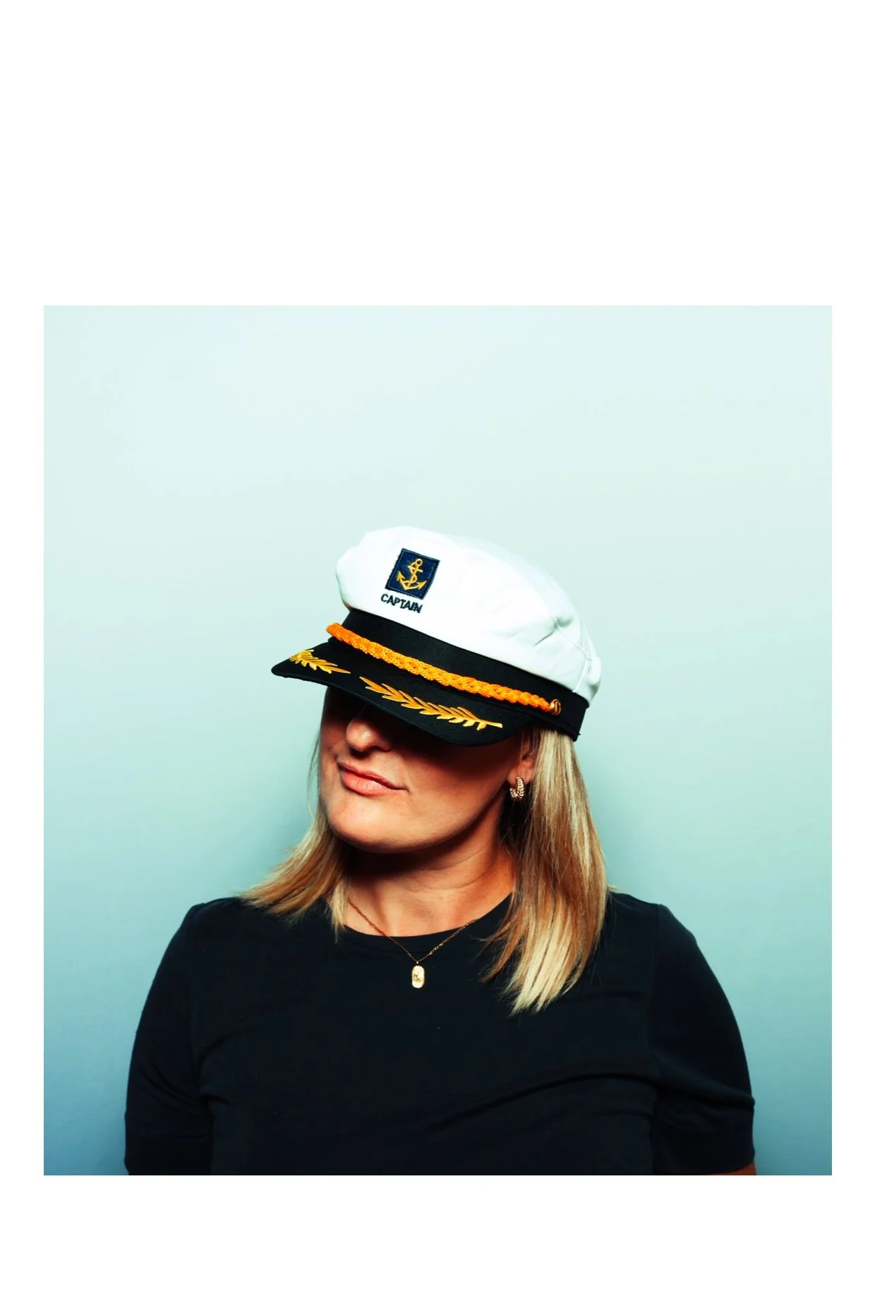 A woman with blonde hair wearing a white captain's hat with a navy blue and gold emblem, a black band, and gold embroidery, a black shirt, and earrings, against a gray background. Taken by Nook booth Photo Booth.