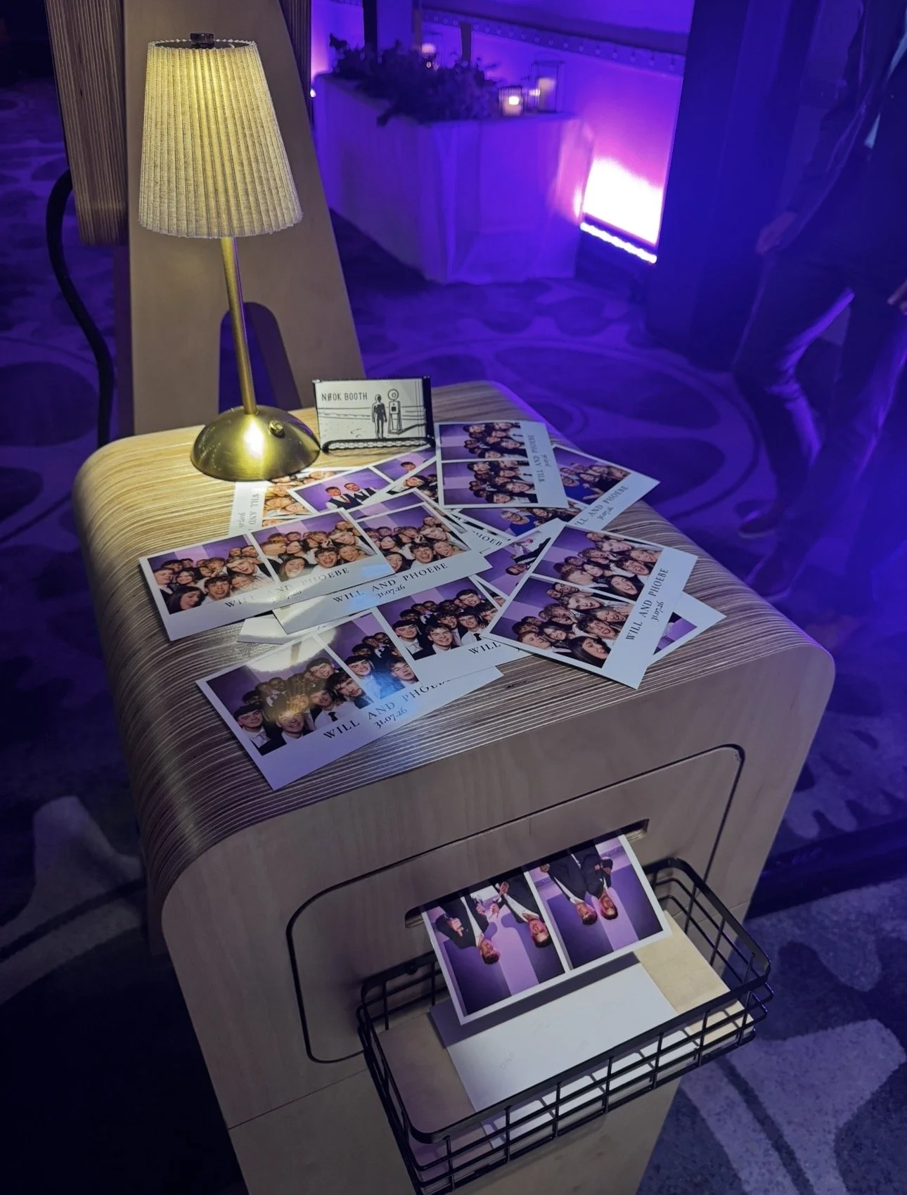 Photograph of a table with numerous photo booth pictures, a small sign indicating a photo booth, a brass table lamp, and candles in the background. The setting is illuminated by purple and yellow lighting, suggesting a festive event or celebration.ti