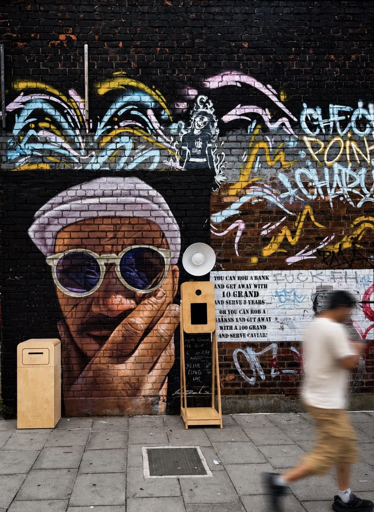 Street art mural of a woman with large sunglasses and a pink hat, holding a finger to her lips, with colorful graffiti and text behind her on a brick wall, and a person walking by in the foreground.