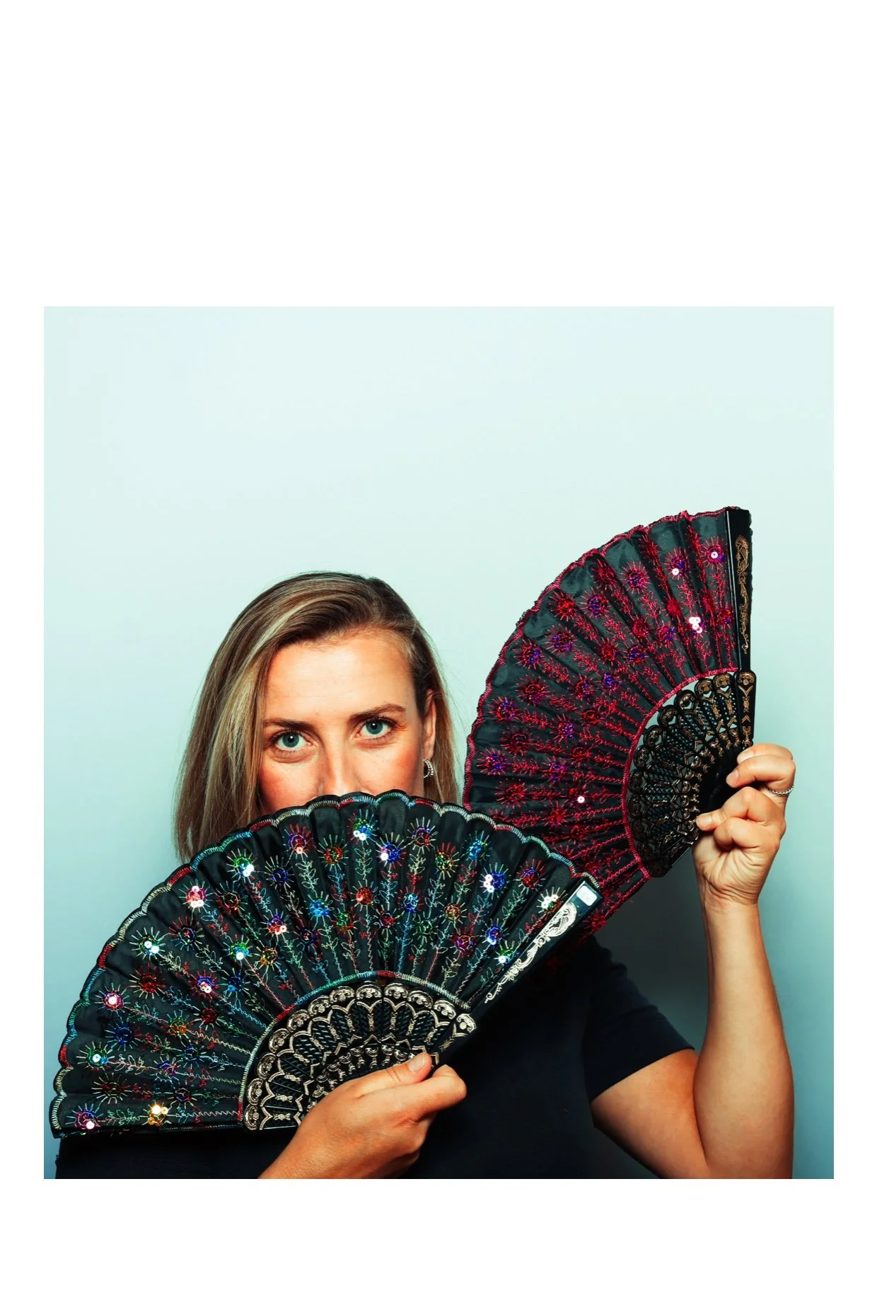 A woman with blonde hair, blue eyes, and hoop earrings holding two decorative black fans with colorful embroidery and sequins, partially covering her face, against a light blue background.